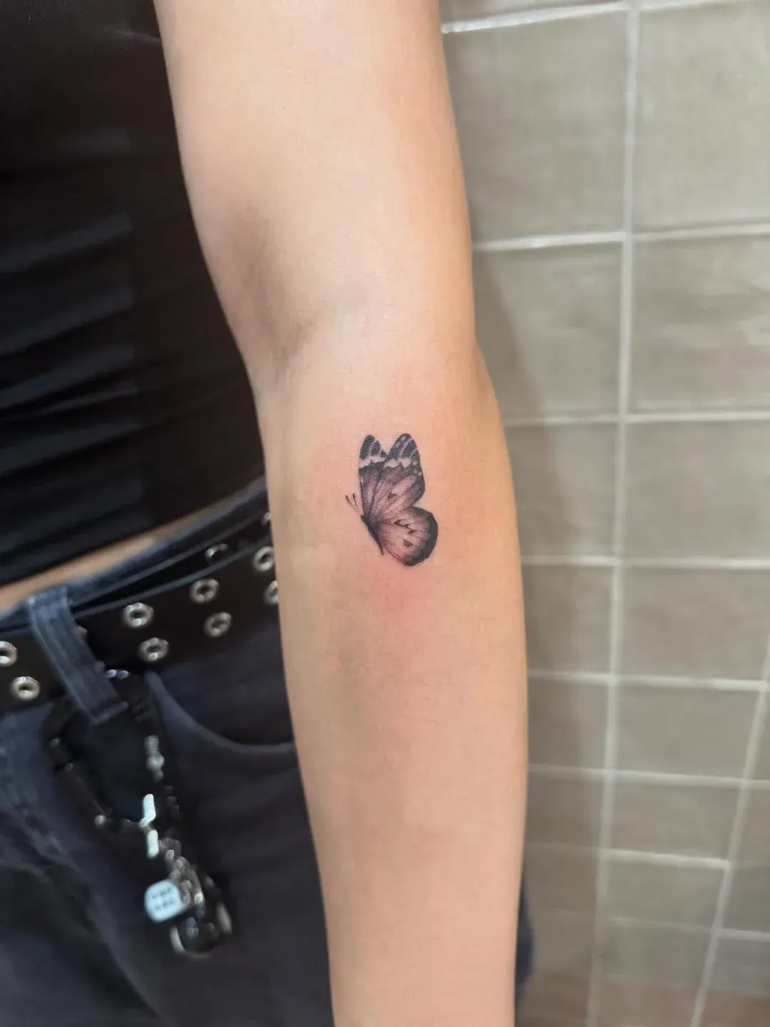 A small, realistic butterfly tattoo on a person's inner forearm against a tiled wall background.