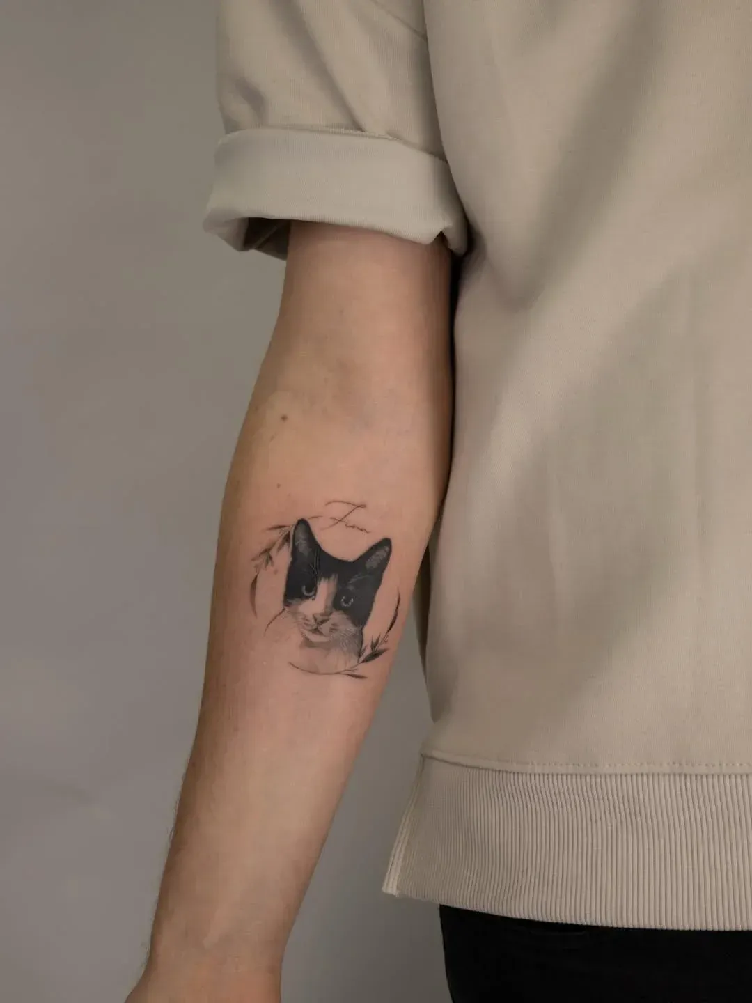 A small, realistic tattoo of a black and white cat's face on the inner forearm of a person wearing a beige shirt.