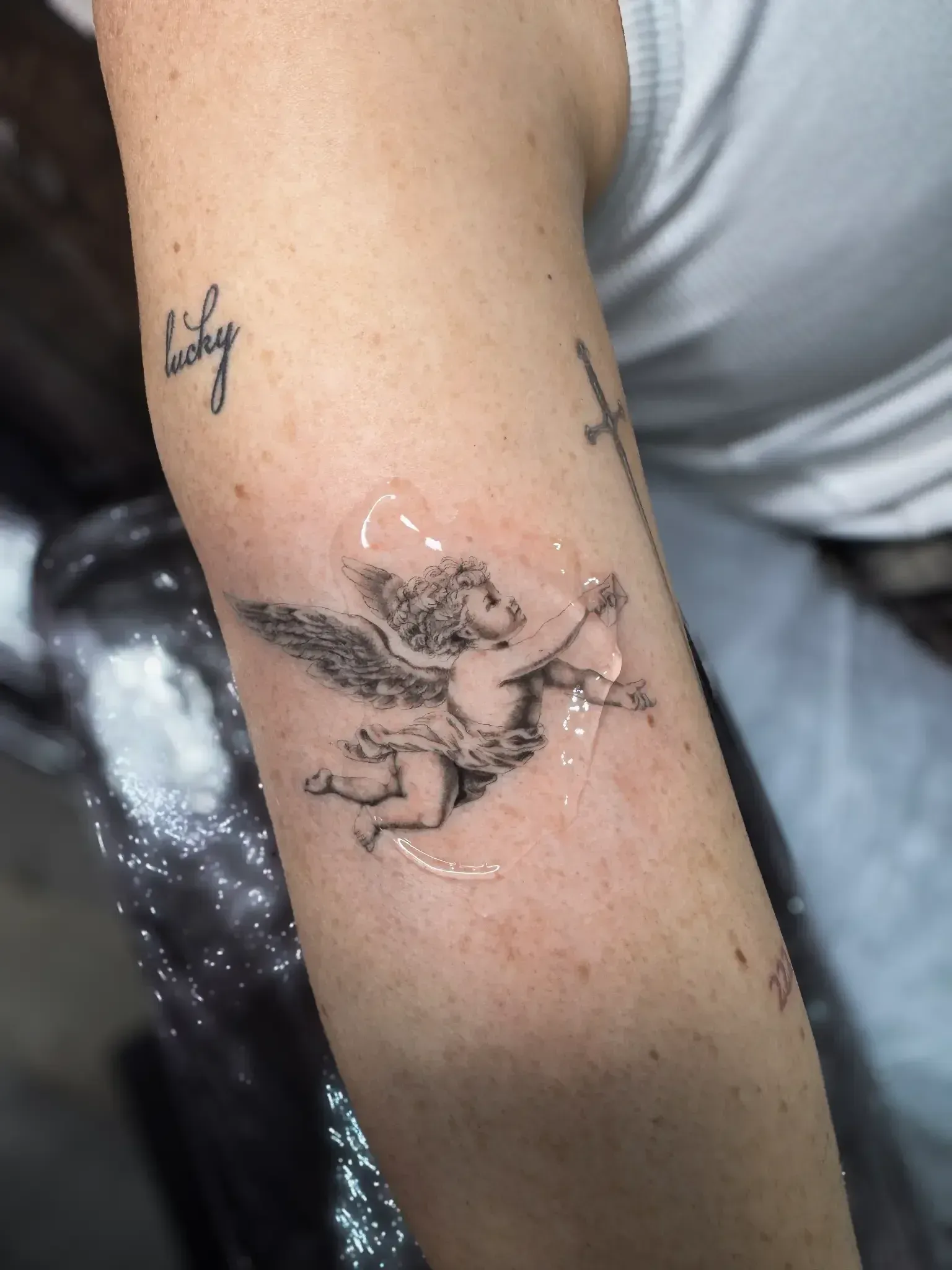 A fresh Cupid tattoo with fine-line shading on a person's upper arm, covered with a thin layer of protective ointment.