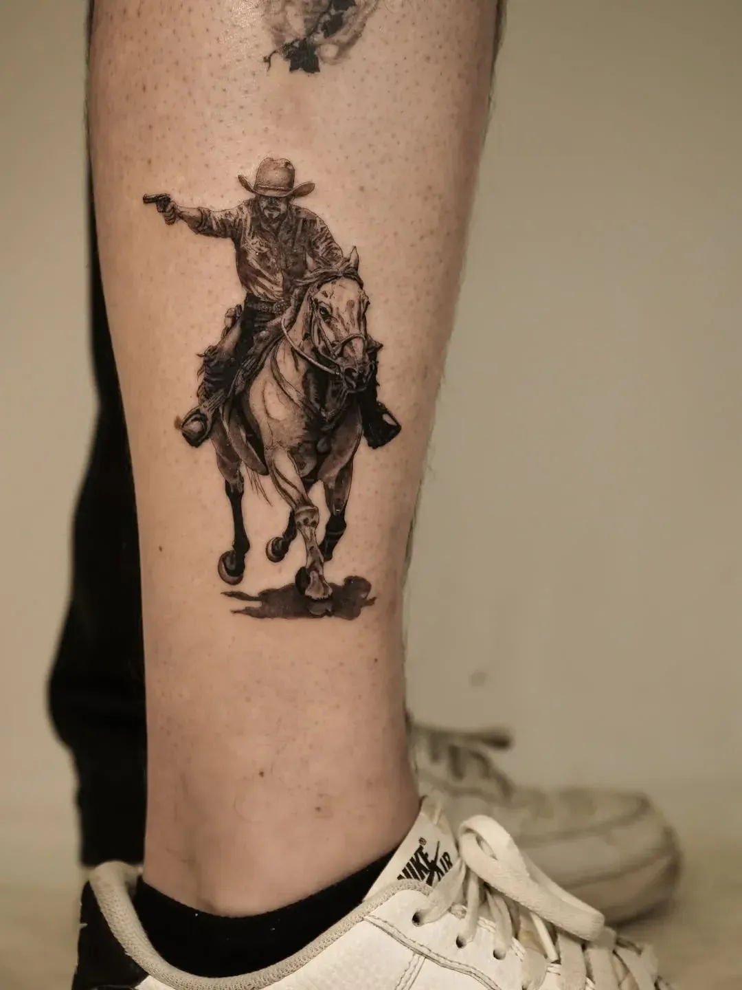 A tattoo of a cowboy riding a galloping horse while pointing a handgun, located on a lower leg.