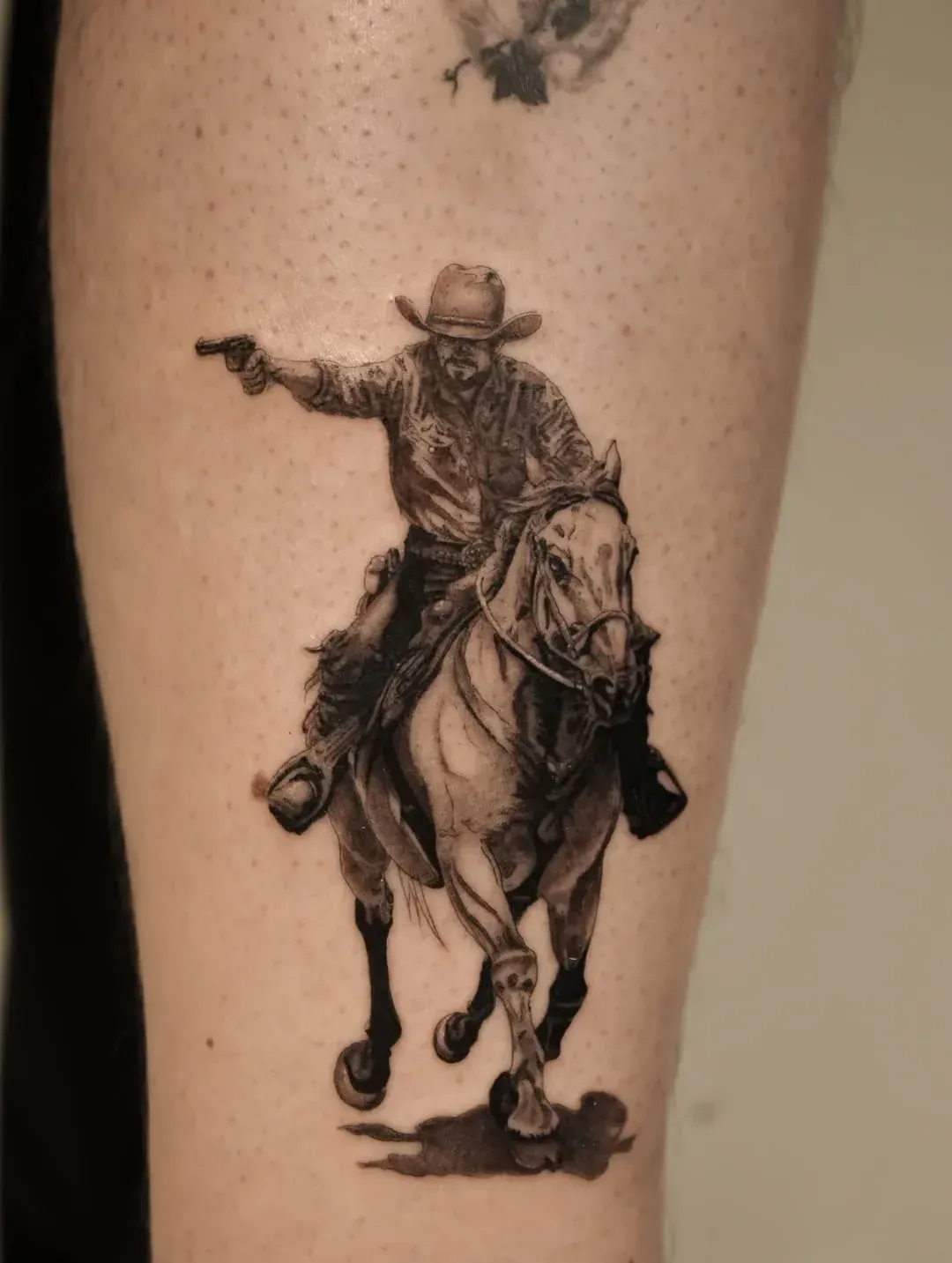 A detailed black-and-grey tattoo of a cowboy riding a horse at a gallop while pointing a pistol to the side.