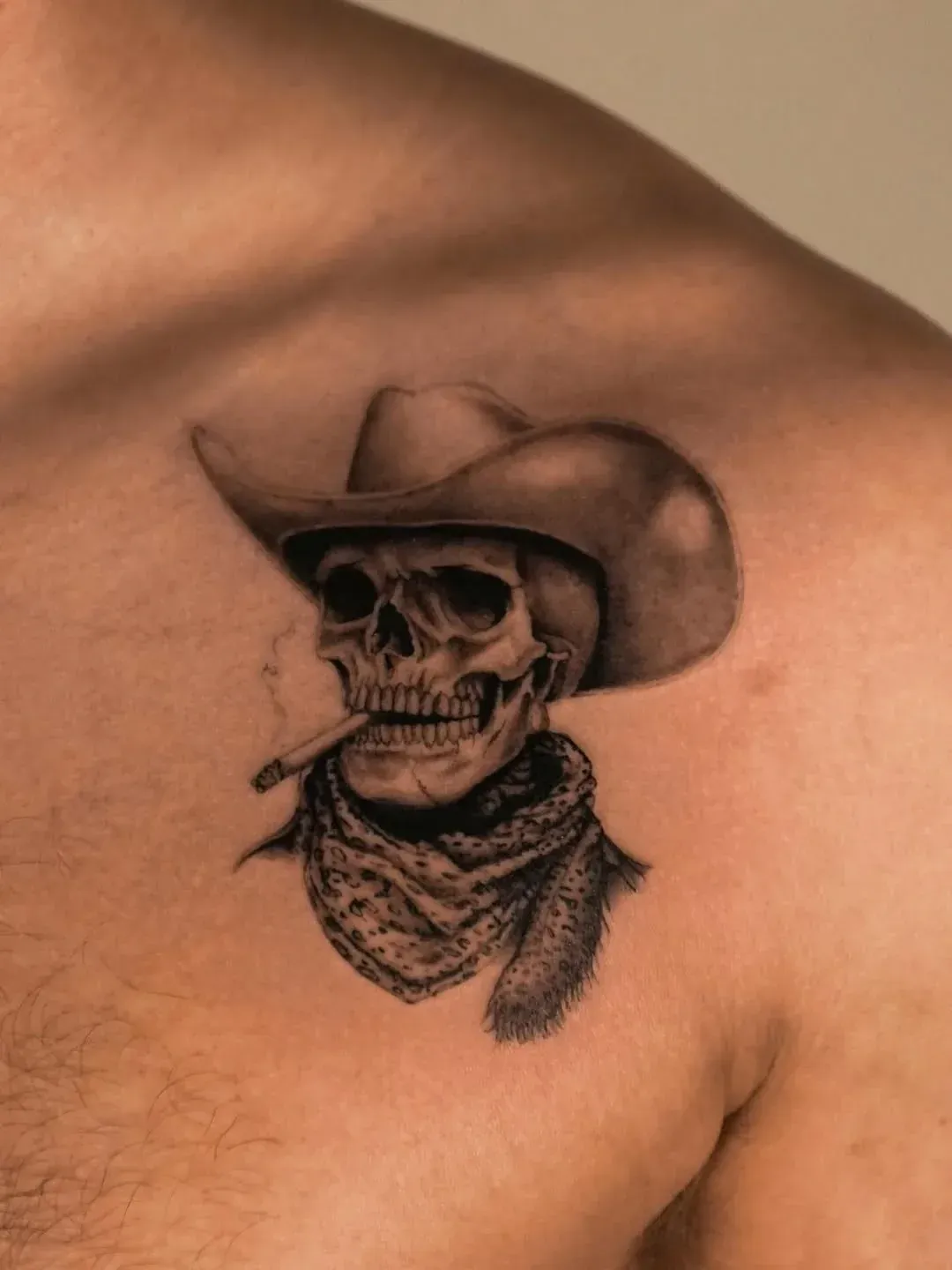 A black and grey tattoo on a person's shoulder depicting a skull wearing a cowboy hat and bandana with a cigarette.