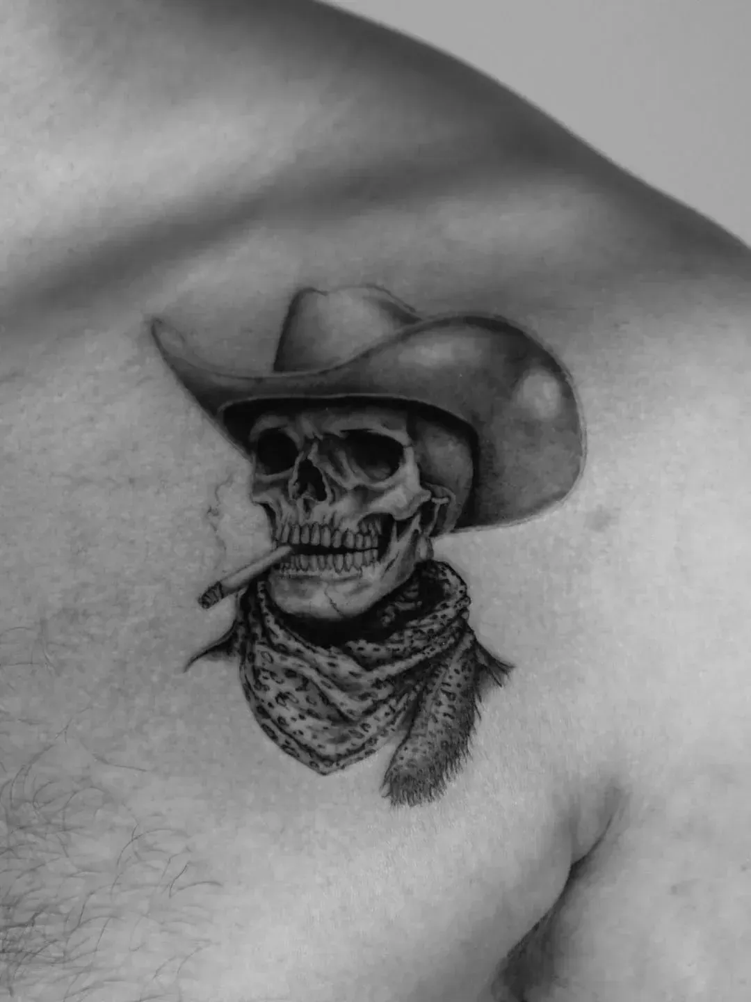 Black-and-white chest tattoo of a skeleton wearing a cowboy hat and patterned bandana with a cigarette in its mouth.