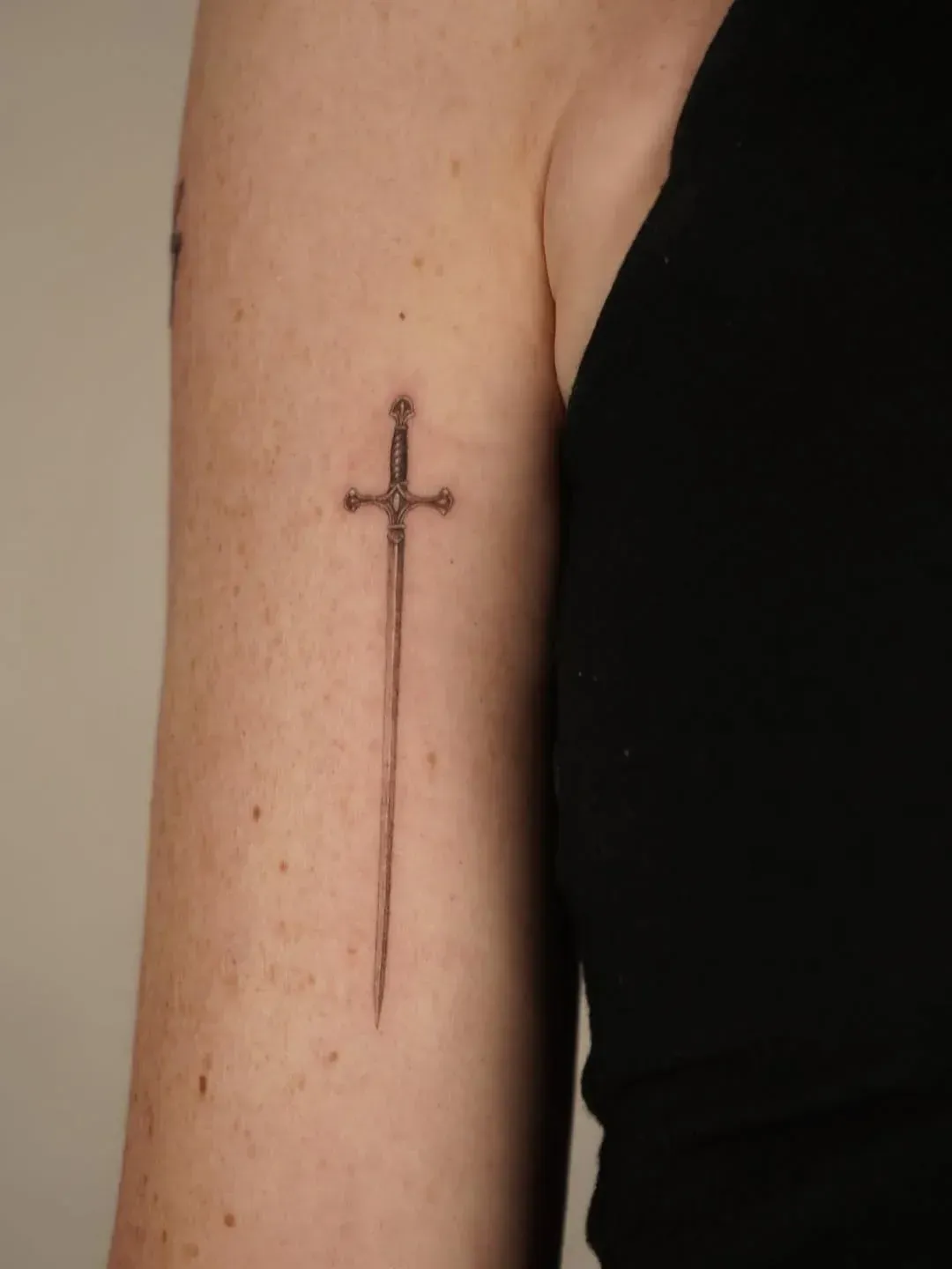 A minimalist, fine-line tattoo of a slender sword centered vertically on the inner upper arm.