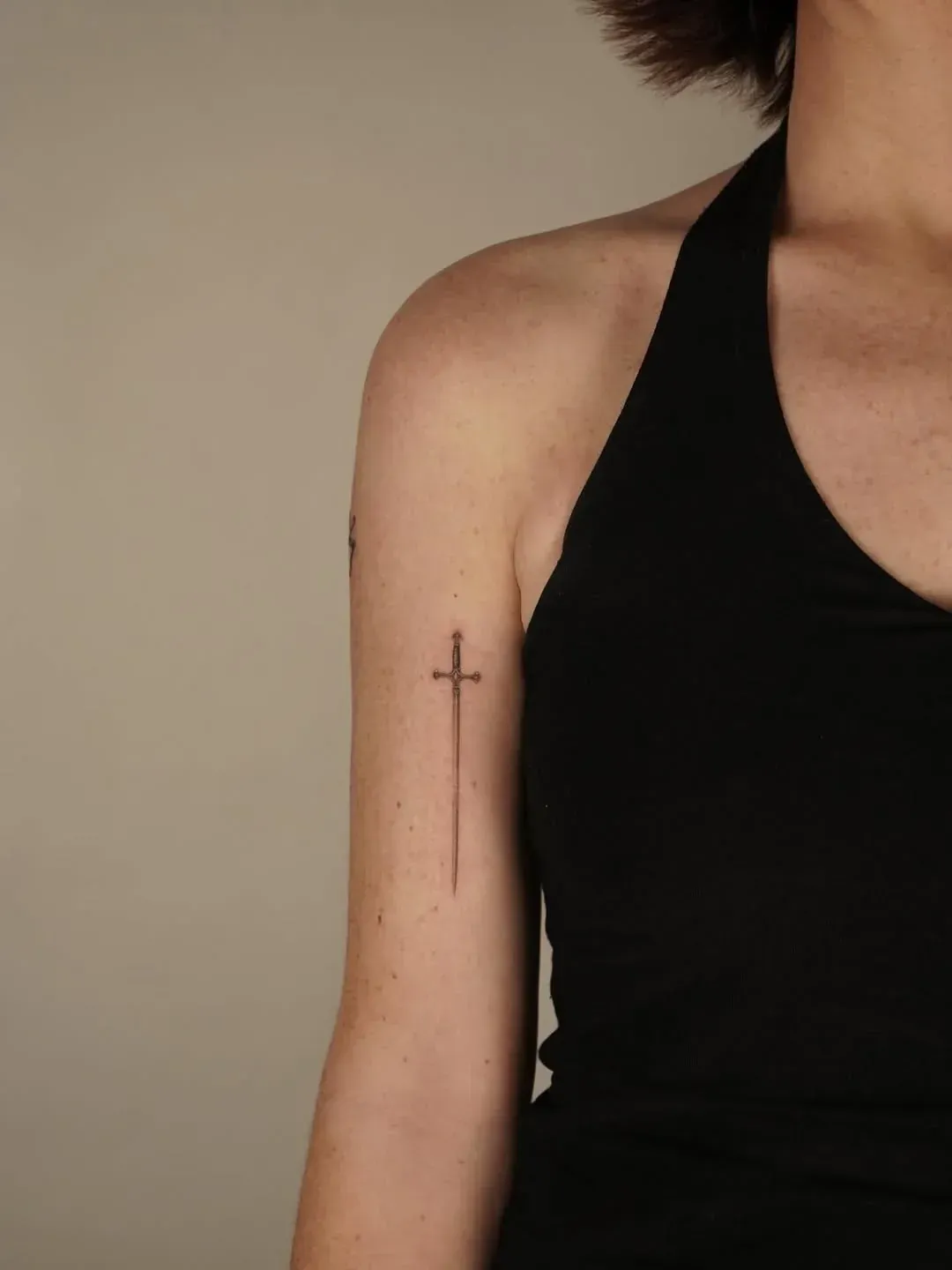 A person wearing a black tank top with a small, fine-line tattoo of a sword on their upper arm.