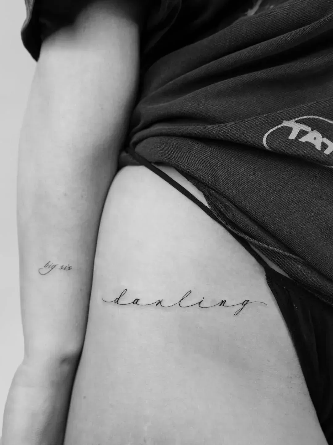 A close-up of a person's torso and arm, featuring delicate script tattoos on the hip and forearm.