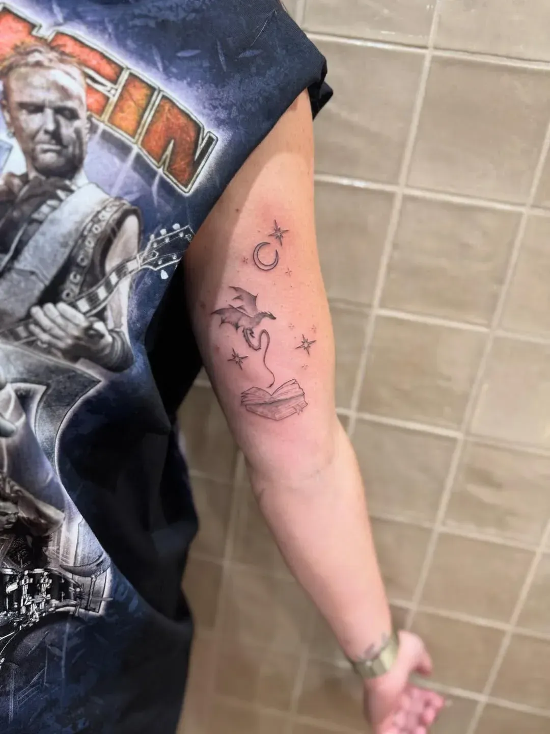 A fine-line tattoo on a person's upper arm featuring a small bat, a moon, stars, and lips.