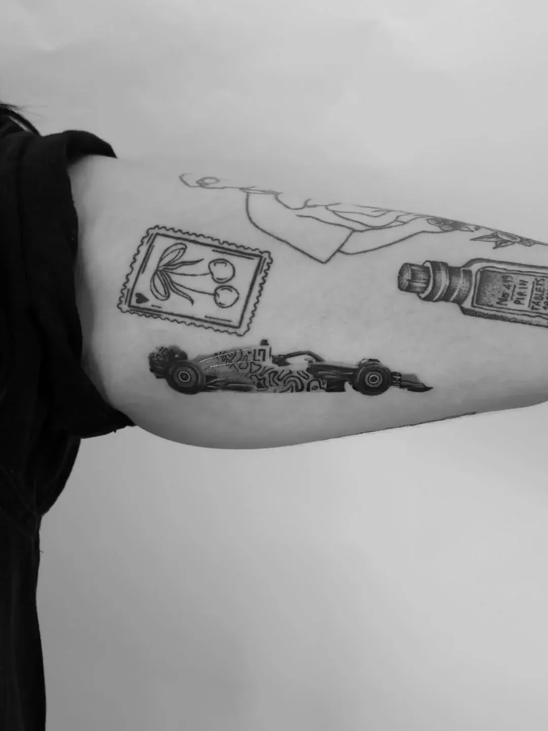 A close-up of a person's upper arm featuring several black ink tattoos, including a racing car, cherries, and a bottle.