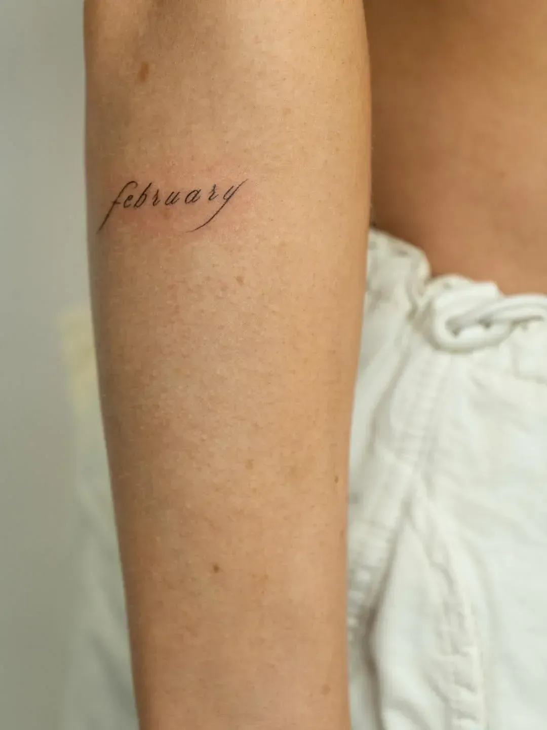 A forearm featuring a minimalist, fine-line cursive tattoo of the word 
