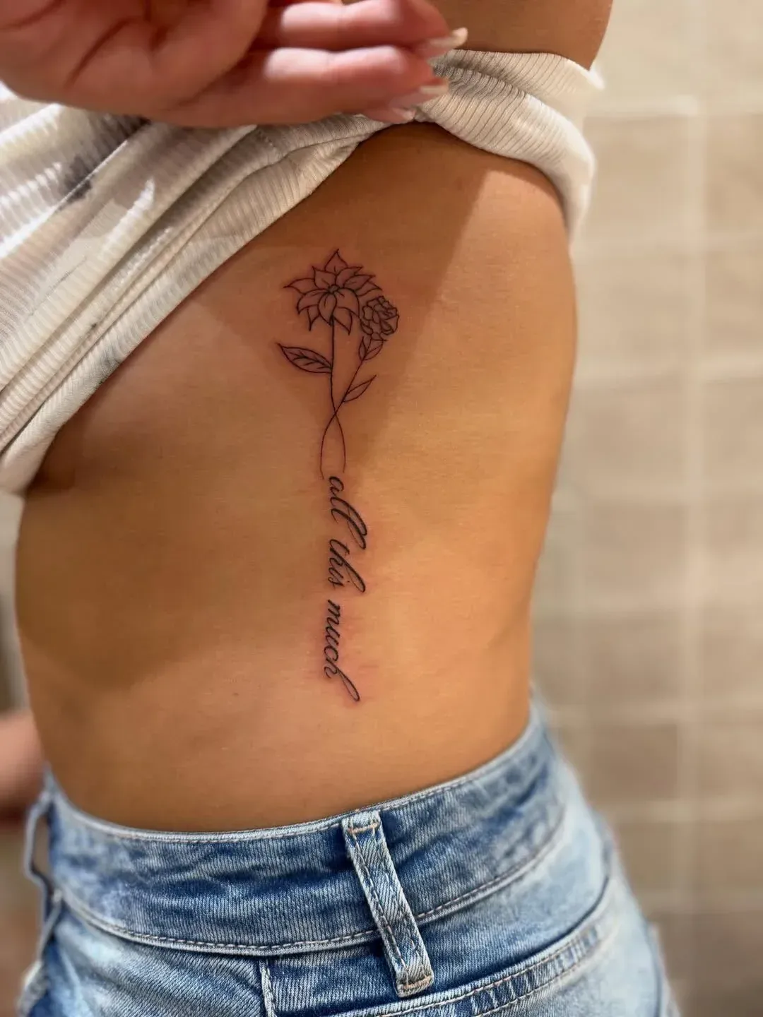 A thin-line tattoo on a person's ribcage featuring a flower with a cursive script stem reading 