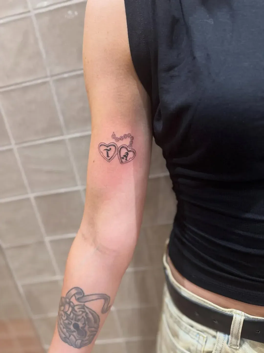A person's arm displaying two small heart tattoos with initials and a larger padlock tattoo on the forearm.