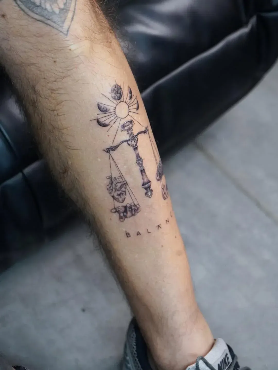 A tattoo on a person's lower leg featuring a scale of justice, a sun with moon phases above it, and the word 