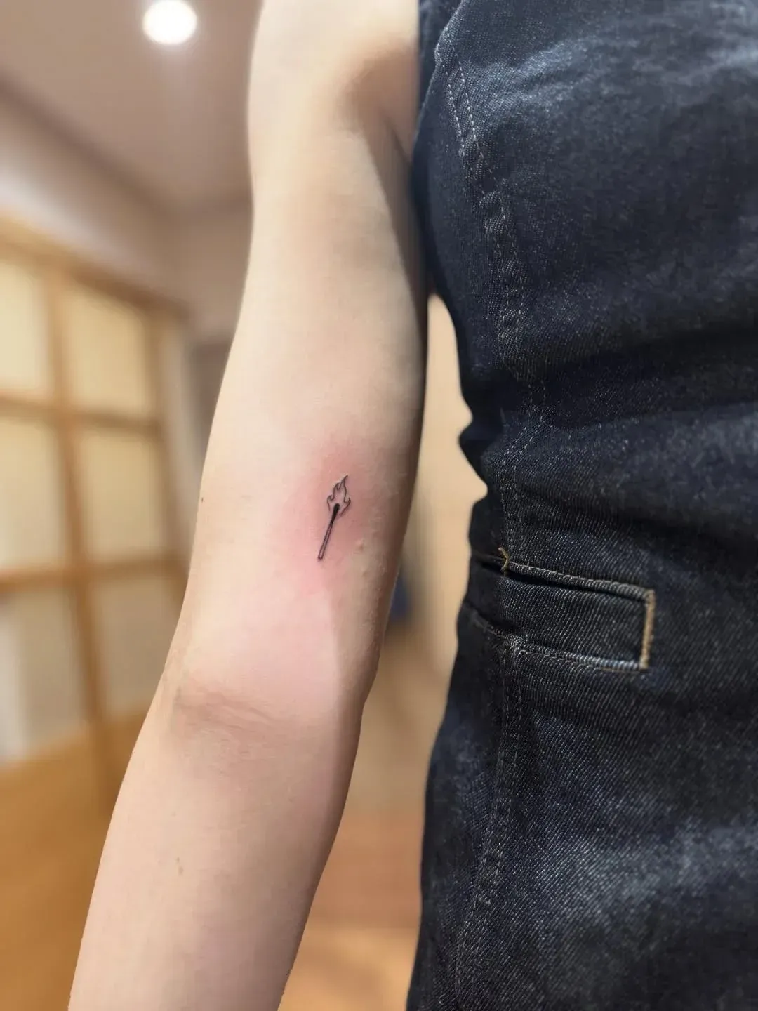 A small, minimalist tattoo of a matchstick on the inner upper arm.