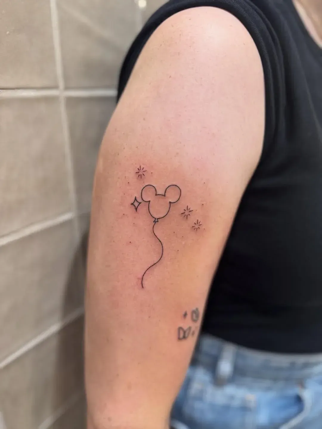 A person's upper arm featuring a fine-line tattoo of a Mickey Mouse-shaped balloon surrounded by small sparkling stars.