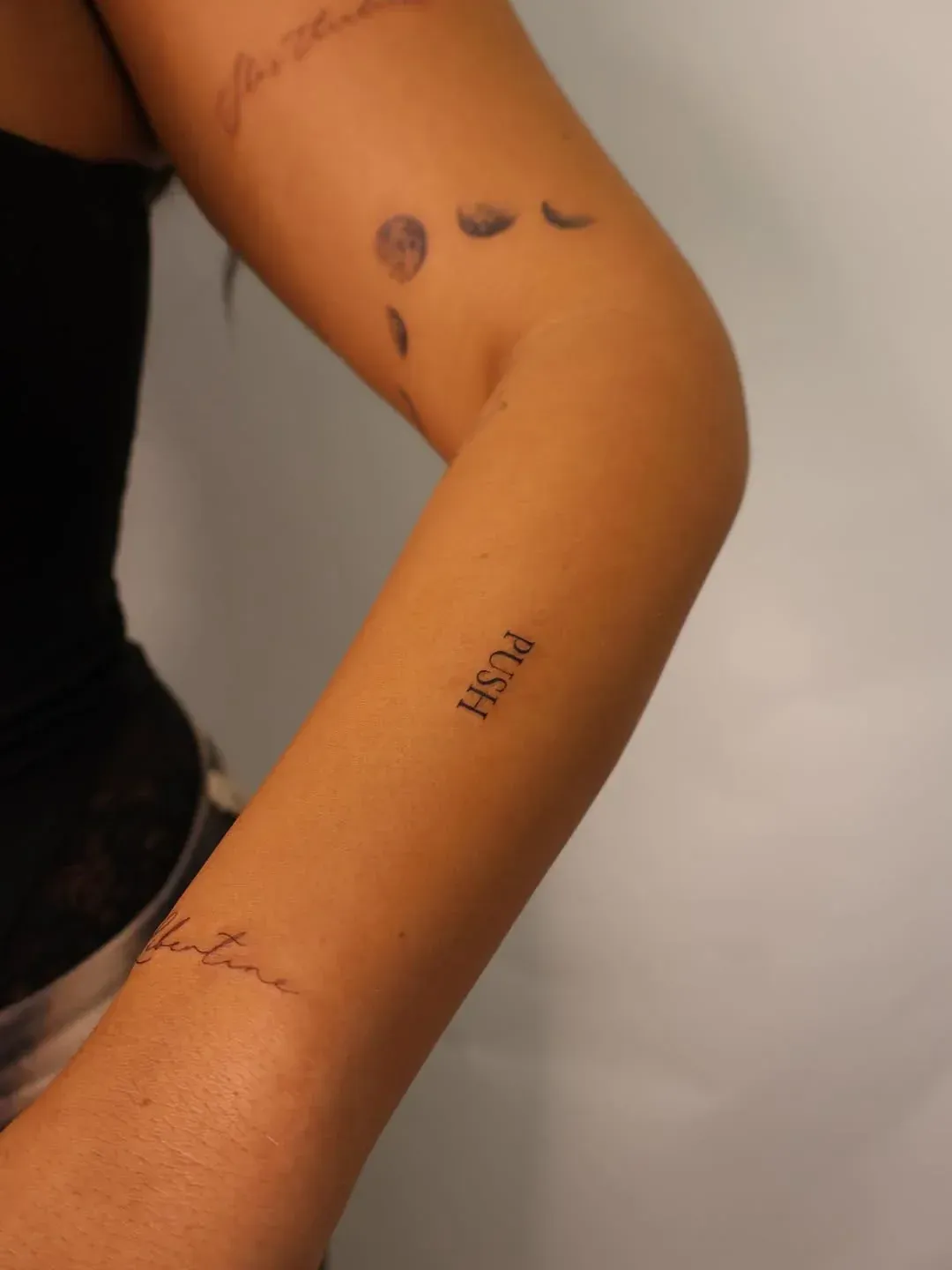 A close-up view of a person's forearm showing three tattoos: a crescent moon phase, the word 