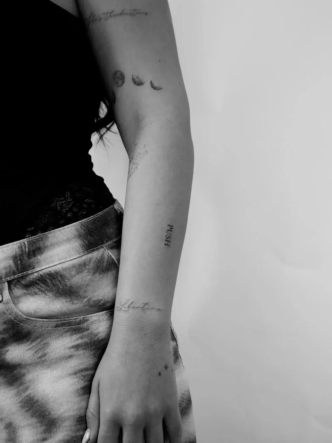 A person’s arm featuring multiple fine-line tattoos, including moon phases, small script text, and a wrist design.