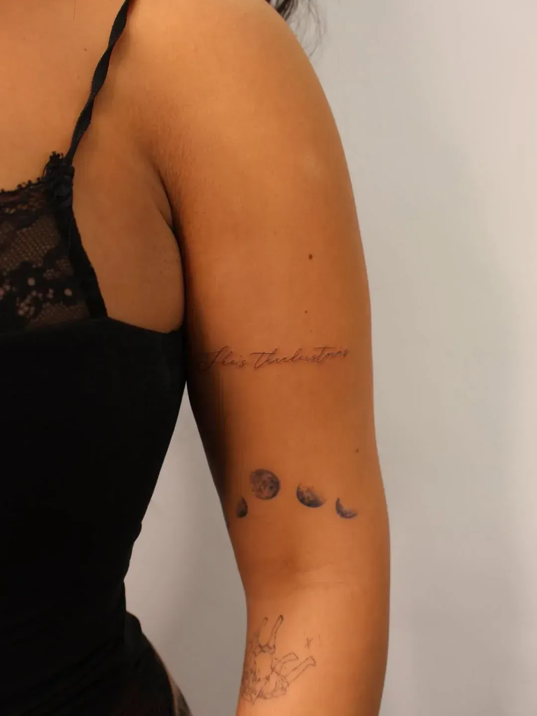 A person's upper arm showing three delicate tattoos: a thin script line, a small moon phase sequence, and a line drawing.