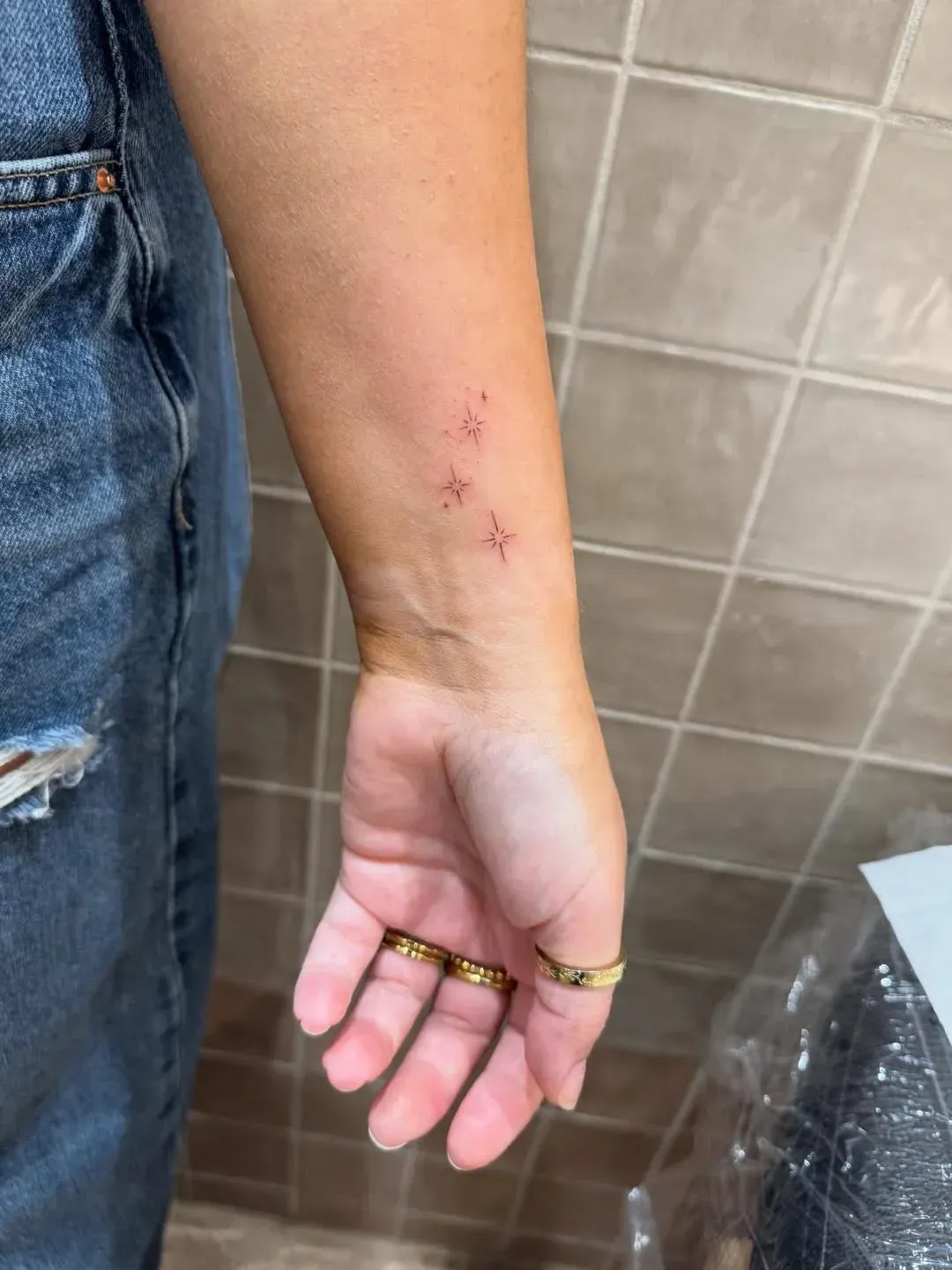 A person's forearm showing three delicate, fine-line, red ink star tattoos against a tiled wall background.
