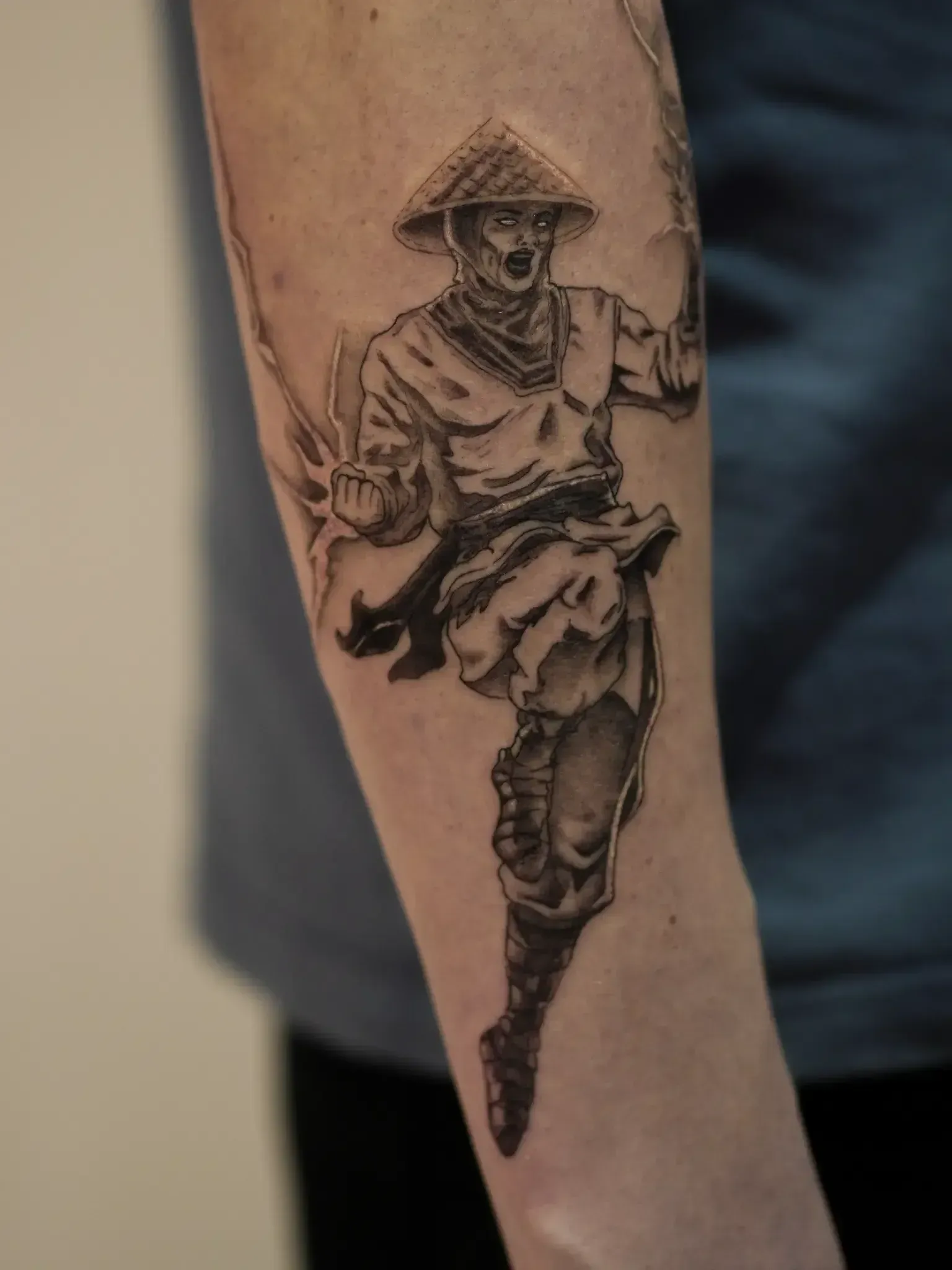 Black and grey tattoo on an arm depicting a dynamic, leaping warrior in a conical hat, mid-action with stylized energy.