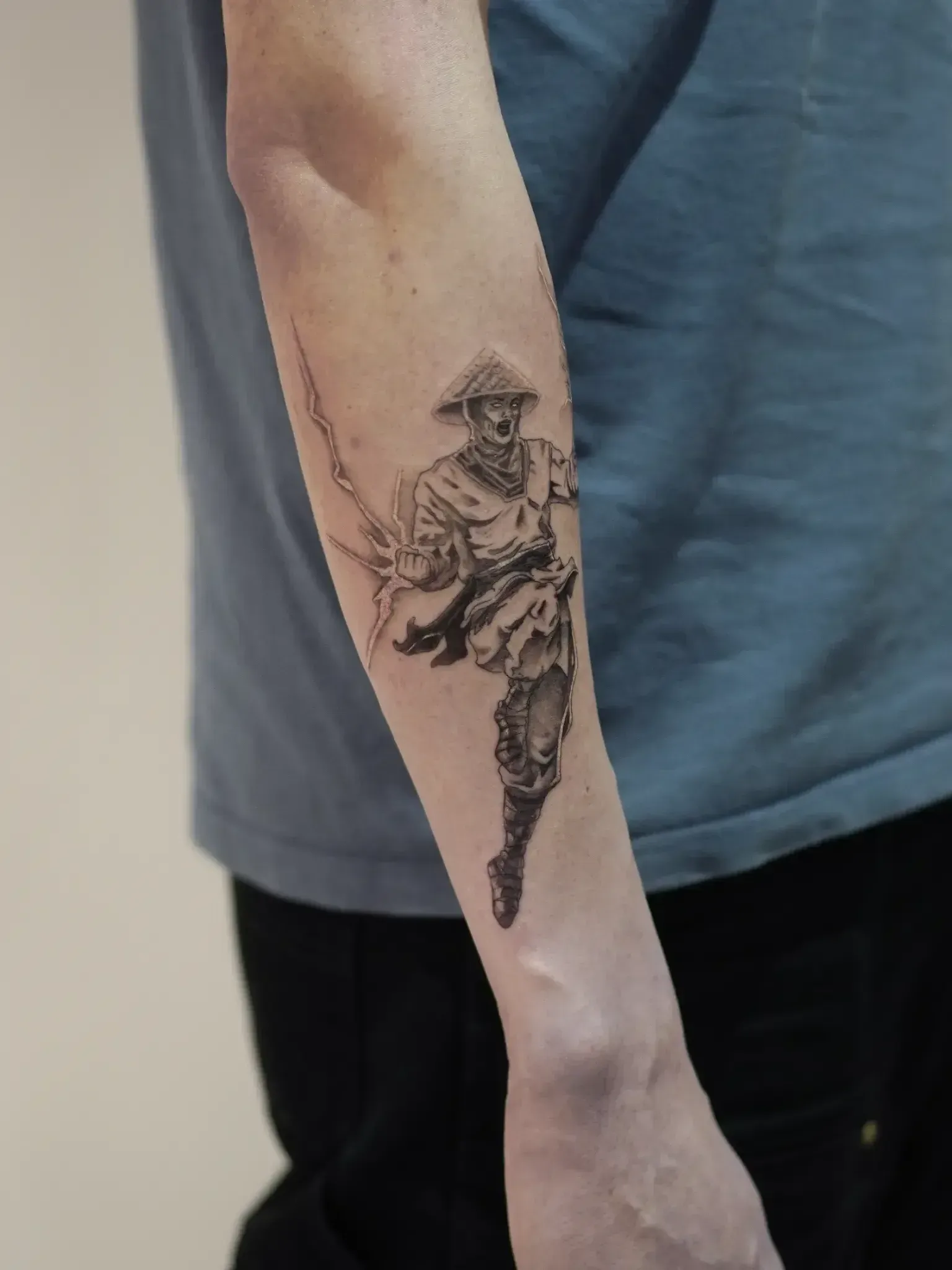 A forearm tattoo showing a stylized warrior in a conical hat, mid-action, with detailed shading and line work.