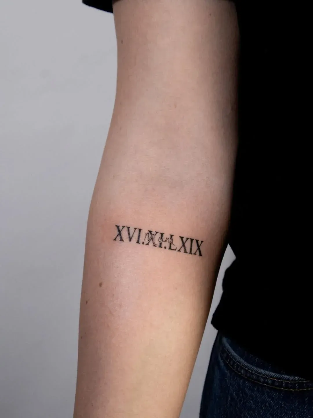 A black ink tattoo on a person's inner forearm featuring the Roman numerals 