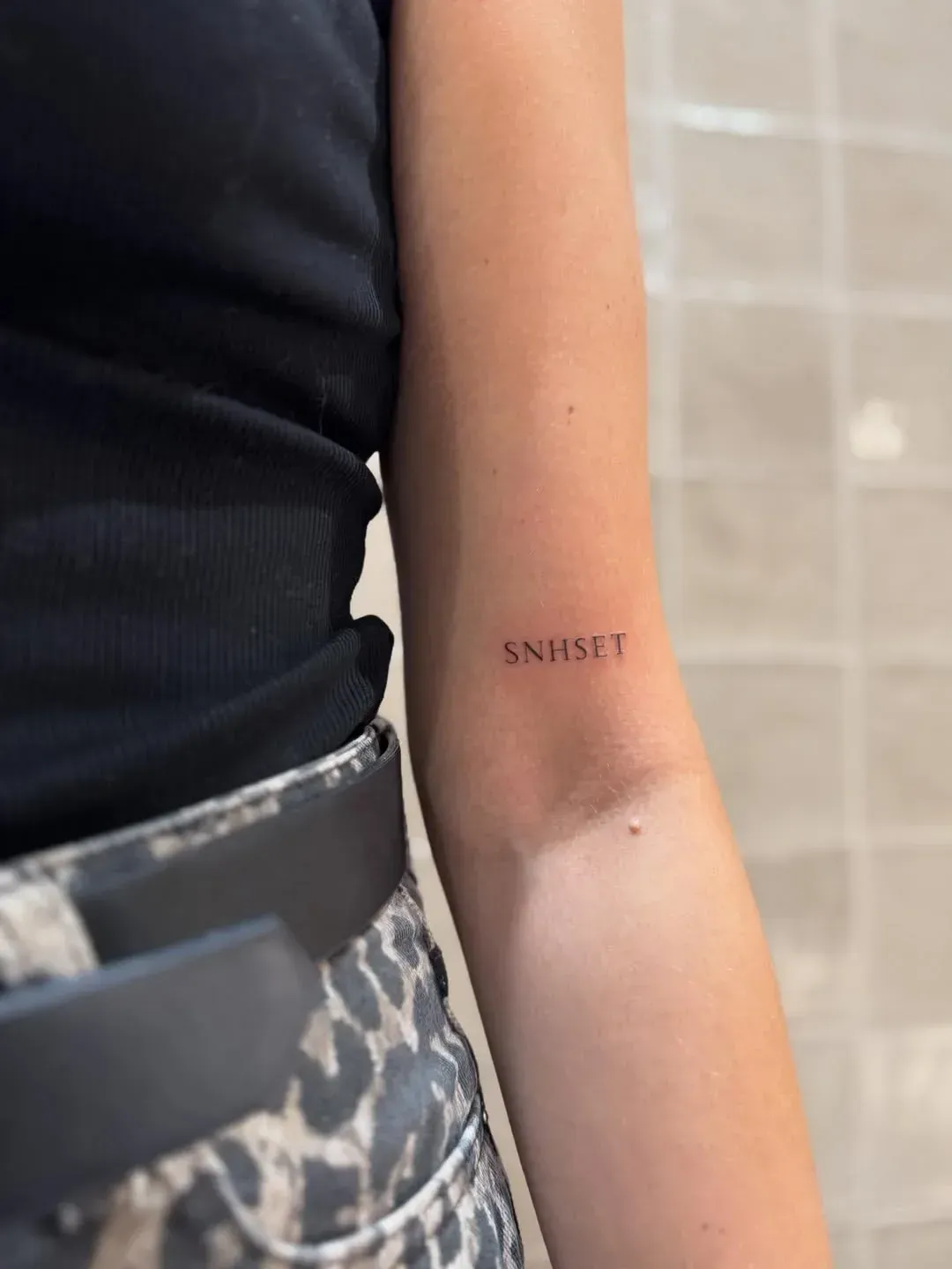A person’s upper arm featuring a small, minimalist tattoo that reads 