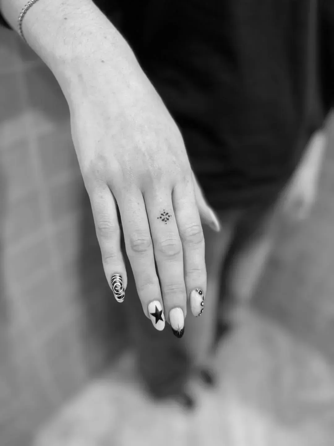 A hand with long, oval nails featuring star and dot art, showcasing a delicate compass tattoo on the middle finger.