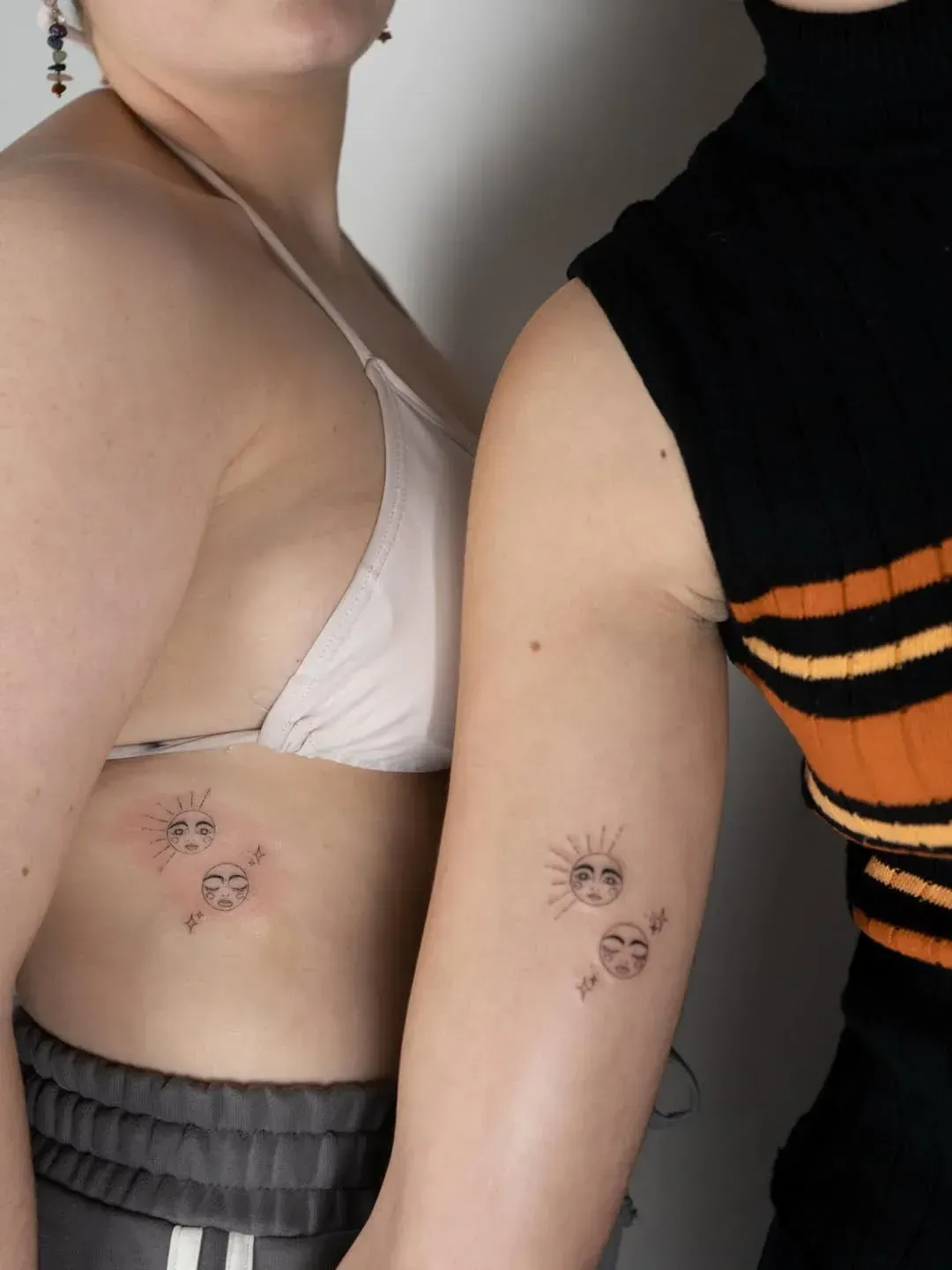 Two people standing side-by-side, each showing identical fine-line tattoos of a sun and a face on their ribcage and arm.