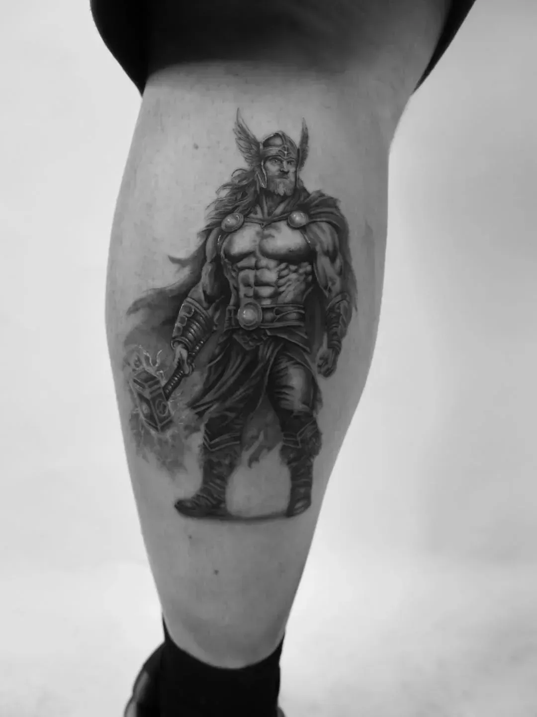Black and white tattoo of a mythical warrior with a winged helmet and a cape, holding a hammer on a person’s lower leg.