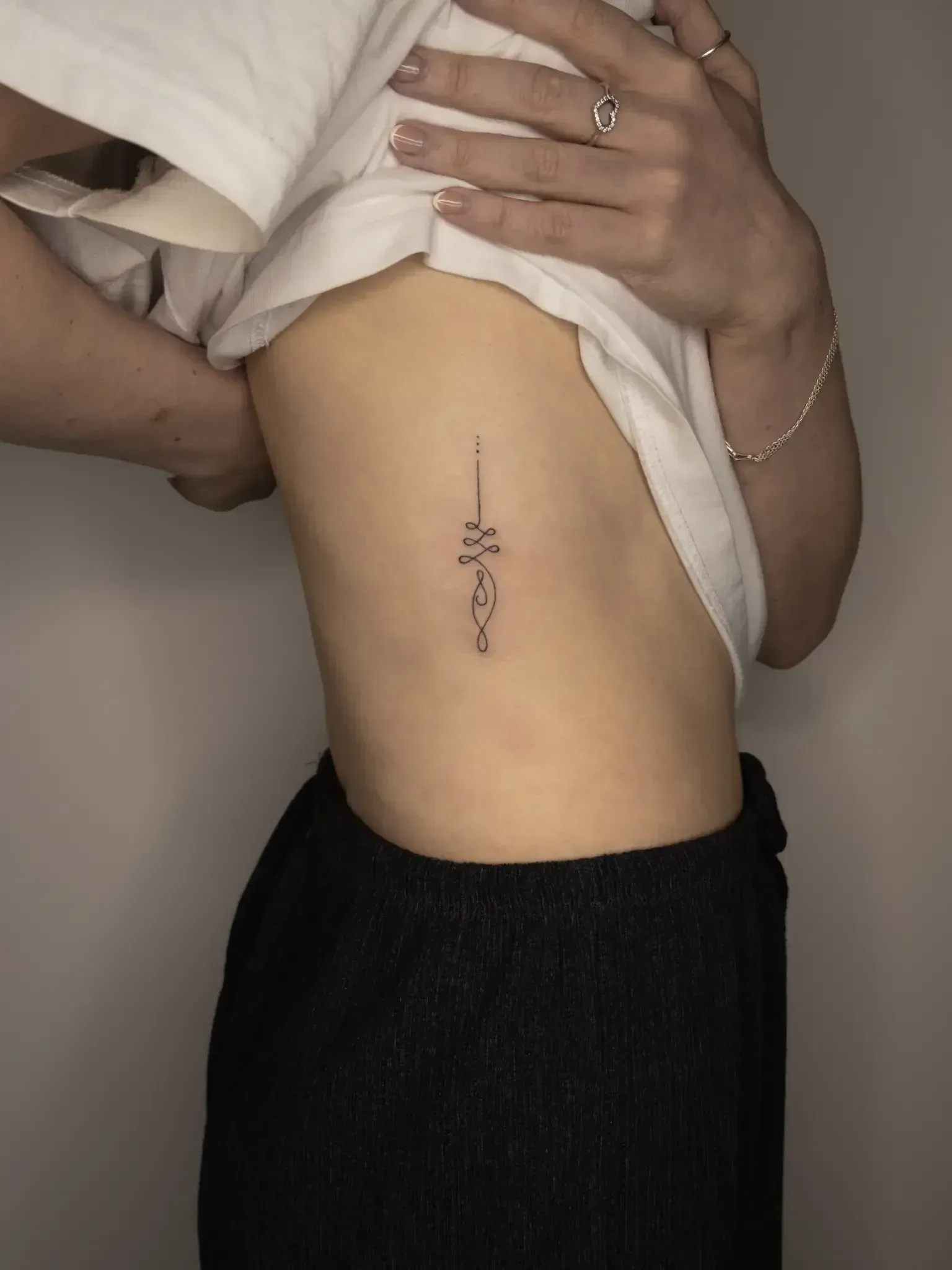 A person showing a small, delicate vertical tattoo on their ribcage featuring a needle and decorative line work.