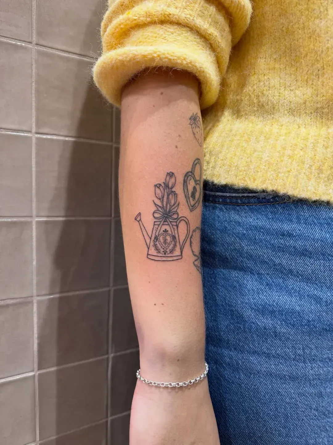 A person wearing a yellow sweater and blue jeans with a fine-line tattoo of a floral watering can on their forearm.