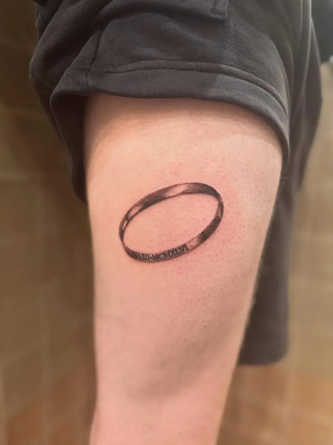 A close-up view of a small tattoo on an arm, depicting a simple, thin brown band or ring with a dark horizontal line.