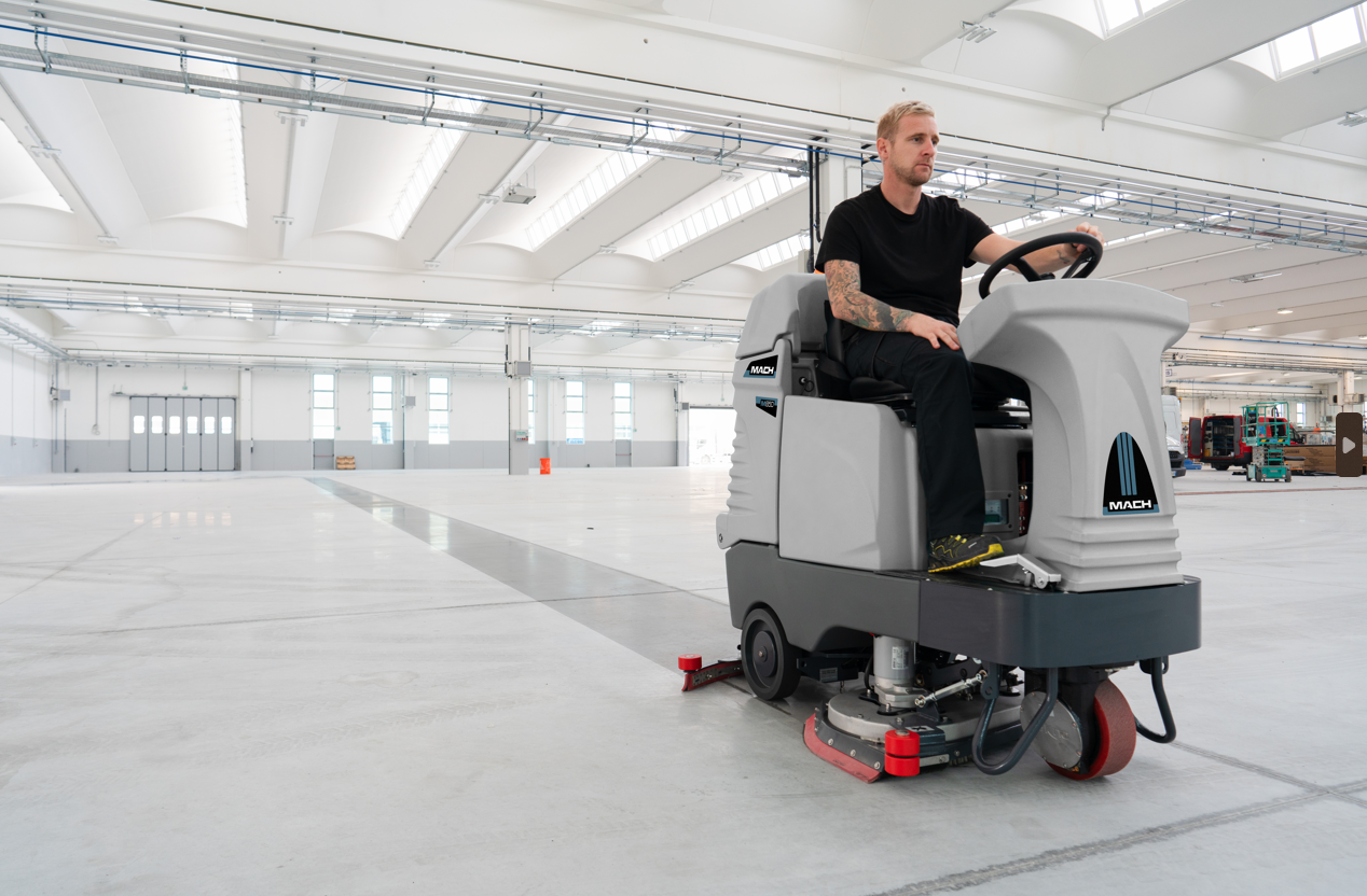 A man is riding a machine on a concrete floor in a warehouse.