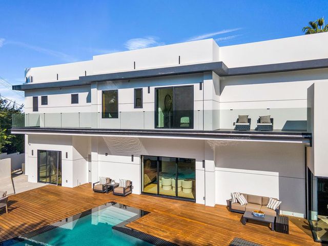 Modern white house with wood deck and pool. Glass balconies, sliding doors, and lounge furniture. Sunny day.