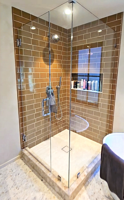 Corner shower with glass doors, brown brick-style tiles, built-in shelf, and marble floor.
