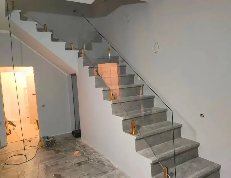 Staircase with gray steps, glass railing, and gold-colored supports, against white walls in a construction setting.