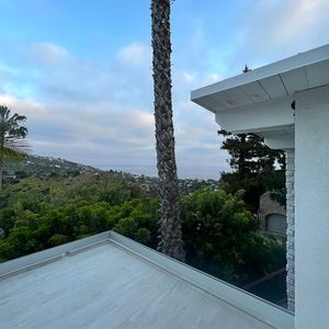Rooftop view of a landscape with green trees, palm tree, and cloudy sky. White building corner in the frame.