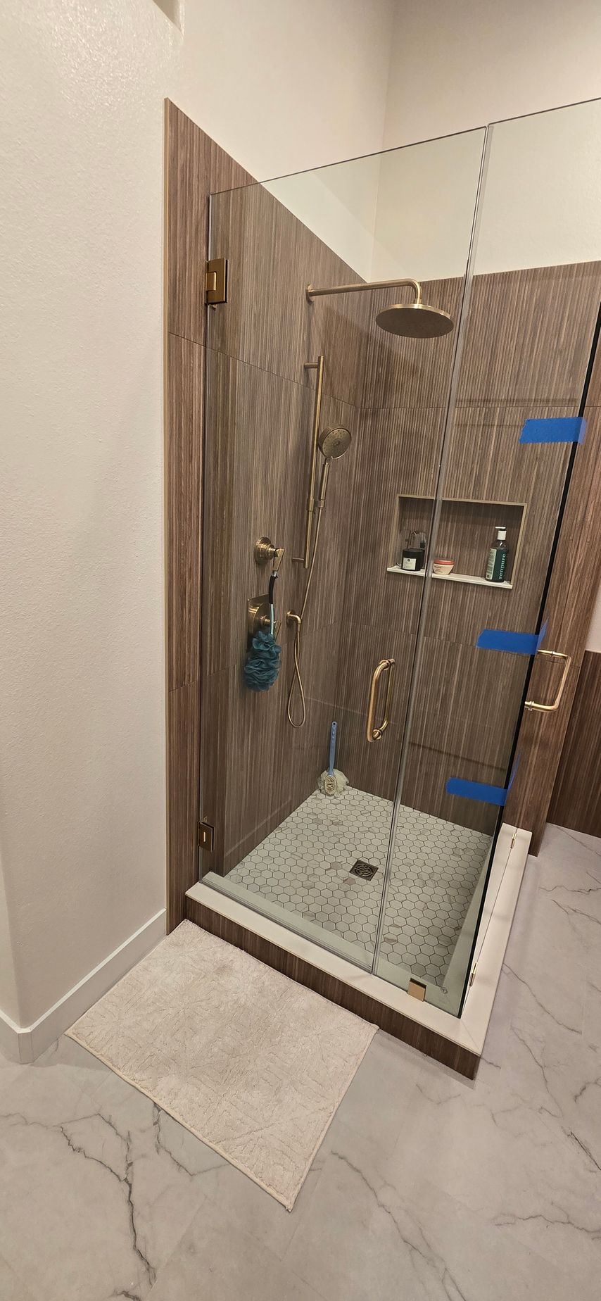Shower stall with brown wood-look tile, glass door, and gold fixtures. White marble floor with light rug.