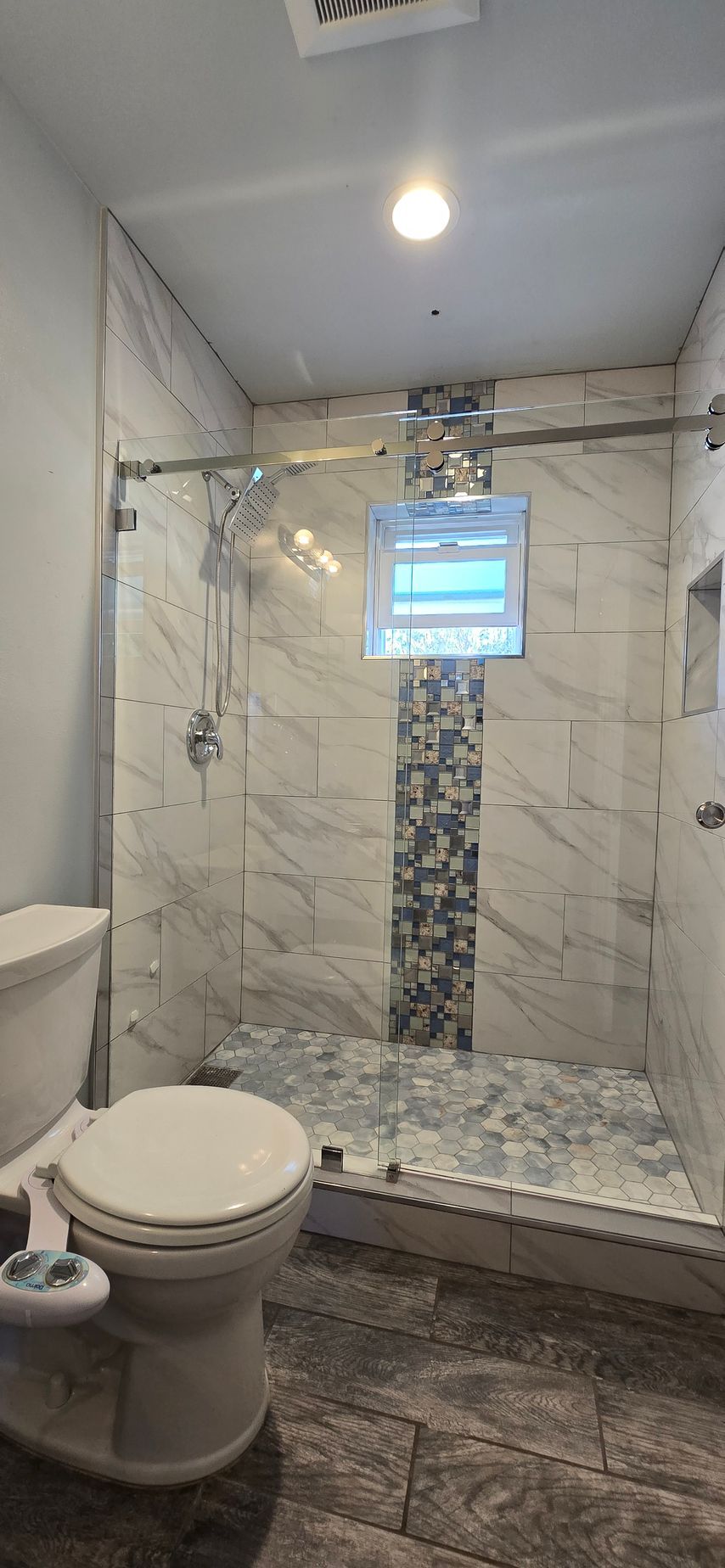 Bathroom with a shower, toilet, and gray tile flooring. The shower has glass doors and marble-like tile.