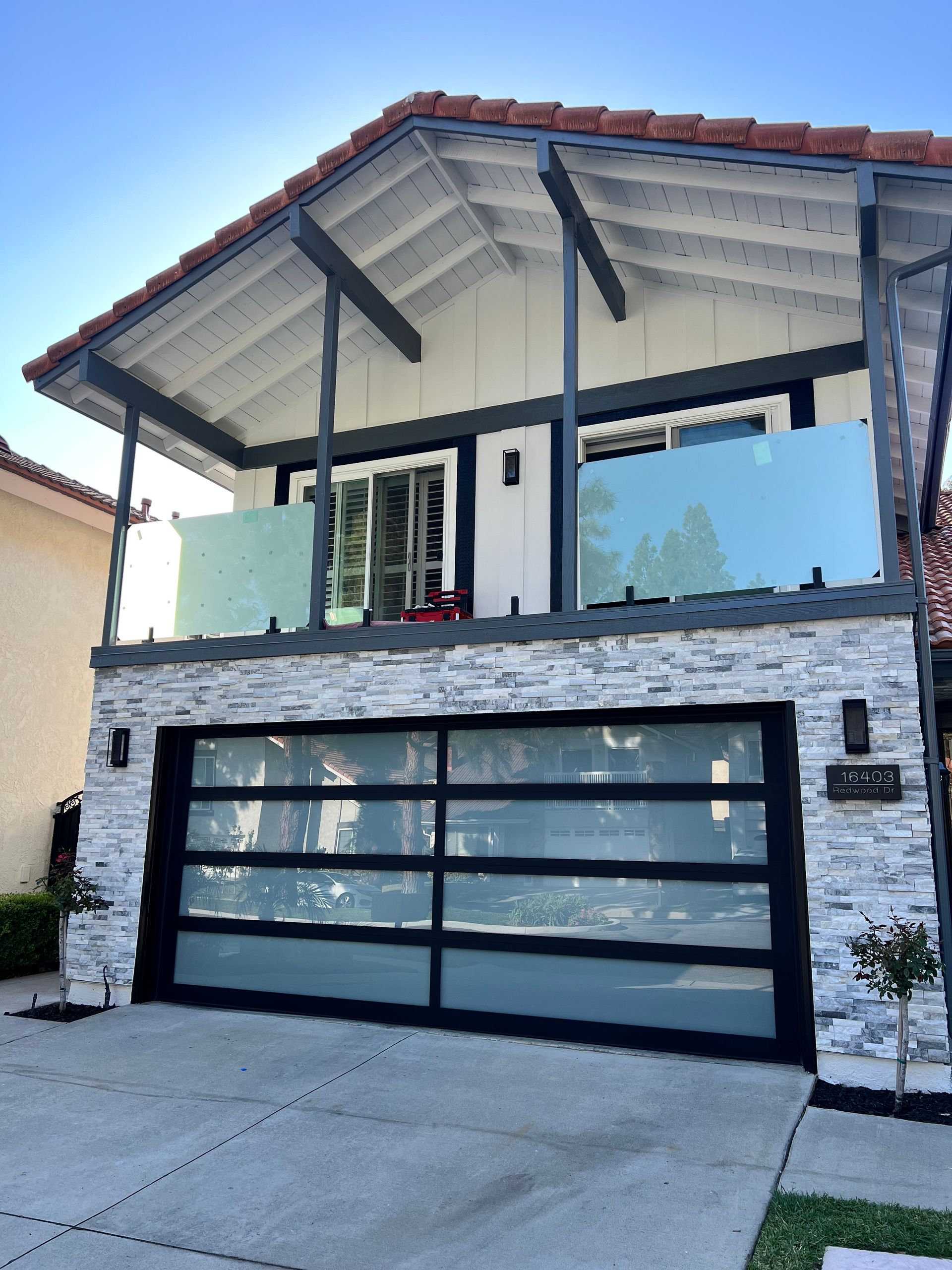 Two-story house with a white and gray facade, glass garage door, and a balcony with glass railings.