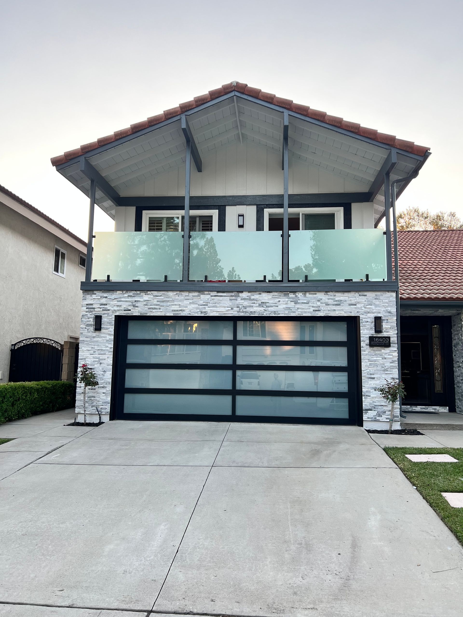 Two-story house with frosted glass garage door and balcony, grey stone facade, and red tile roof.