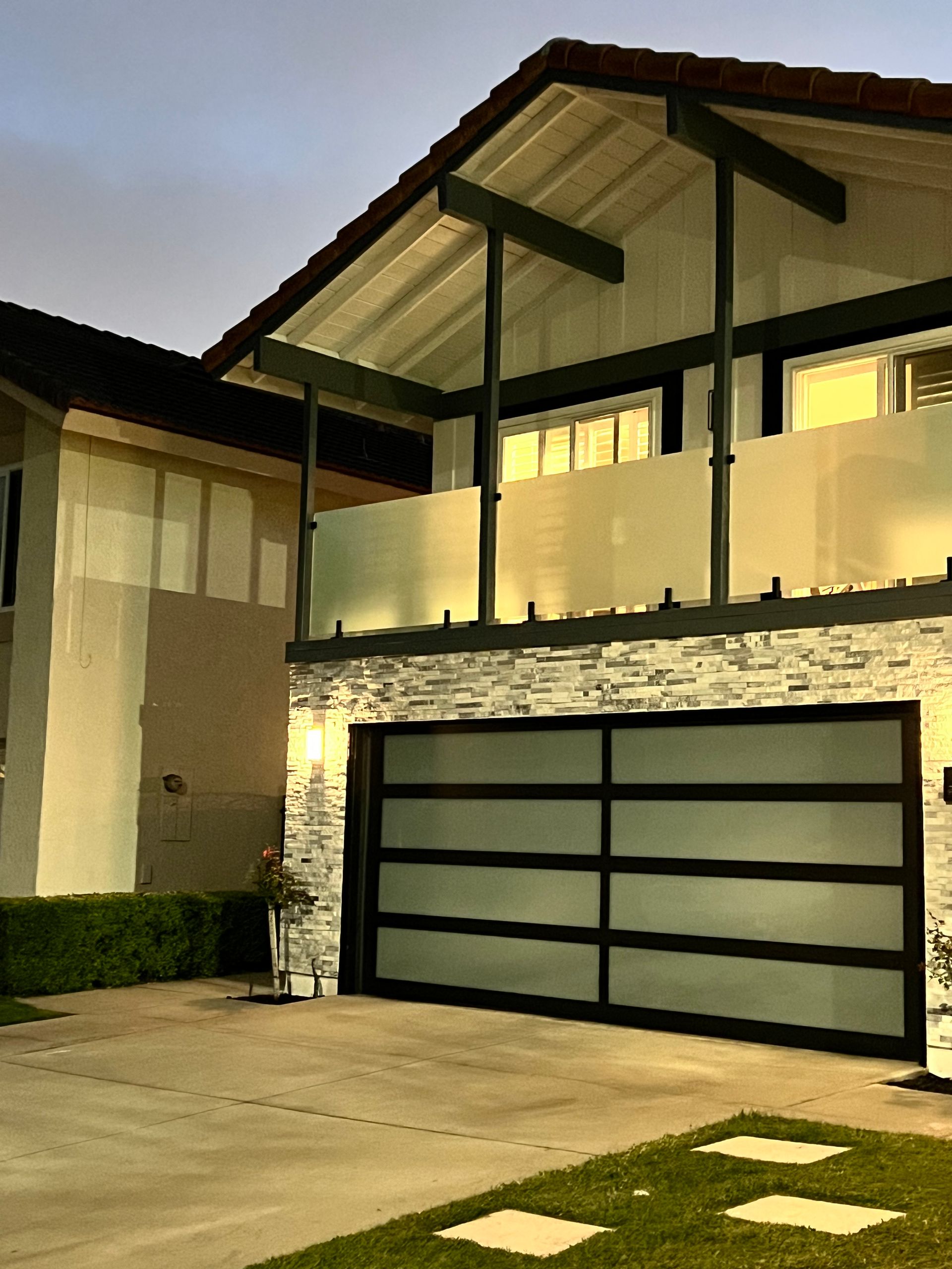 Modern house exterior with stone facade, glass garage door, and second-story balcony under a cloudy sky.