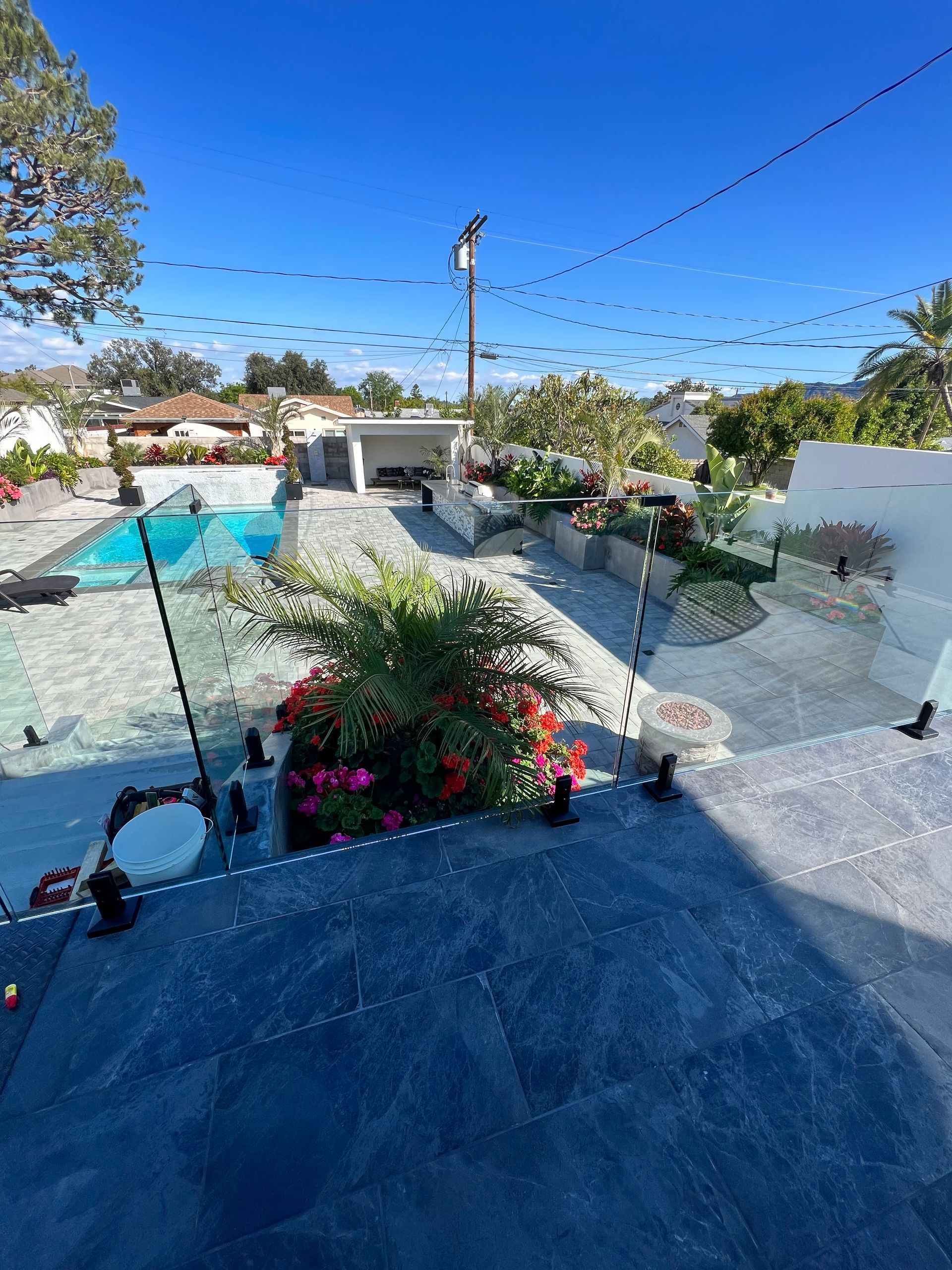 Backyard with blue pool, gray stone patio, and vibrant flower beds under a bright blue sky.