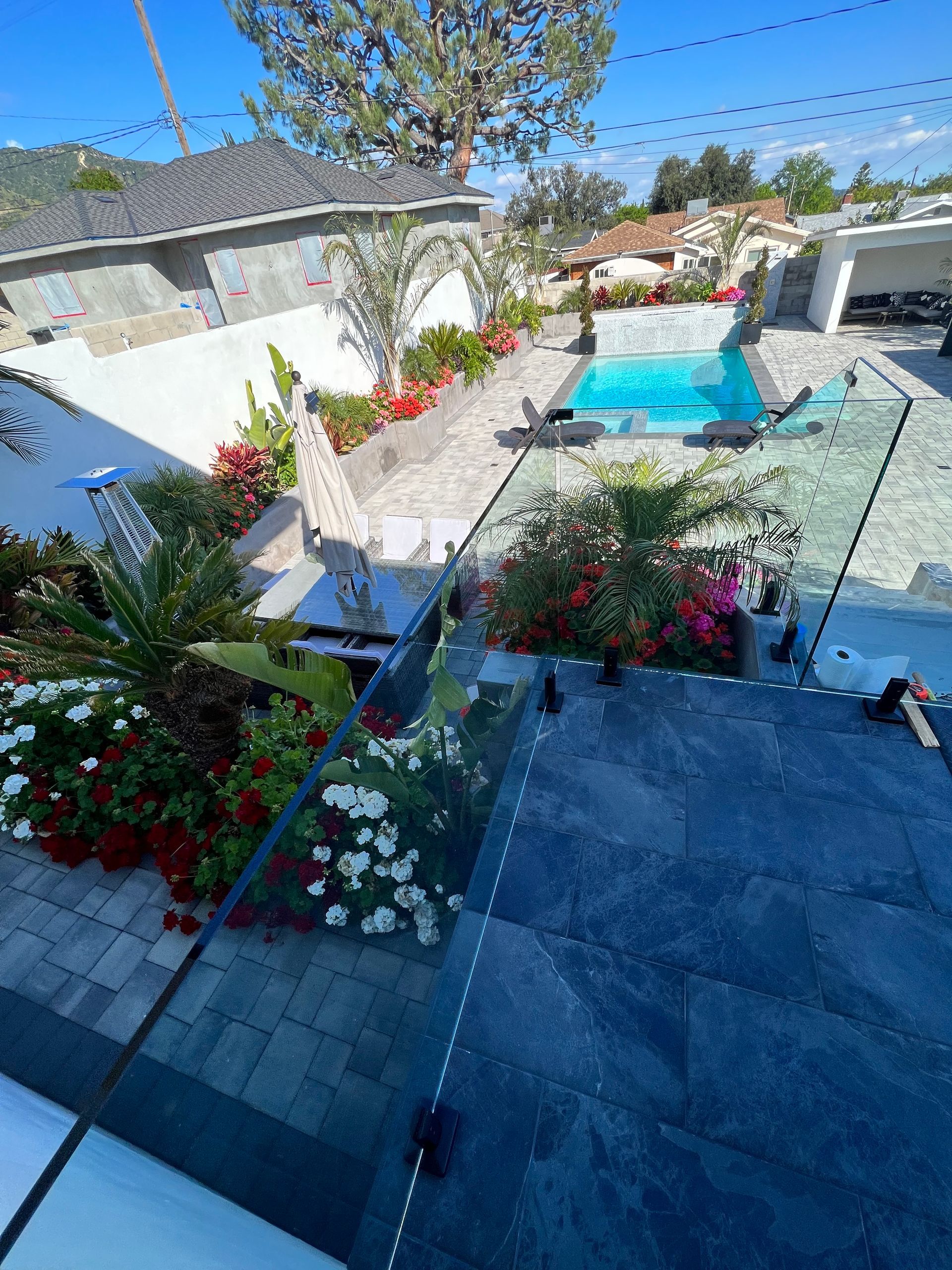 Backyard with pool, flowers, patio, and glass railing. Blue water and a sunny day.
