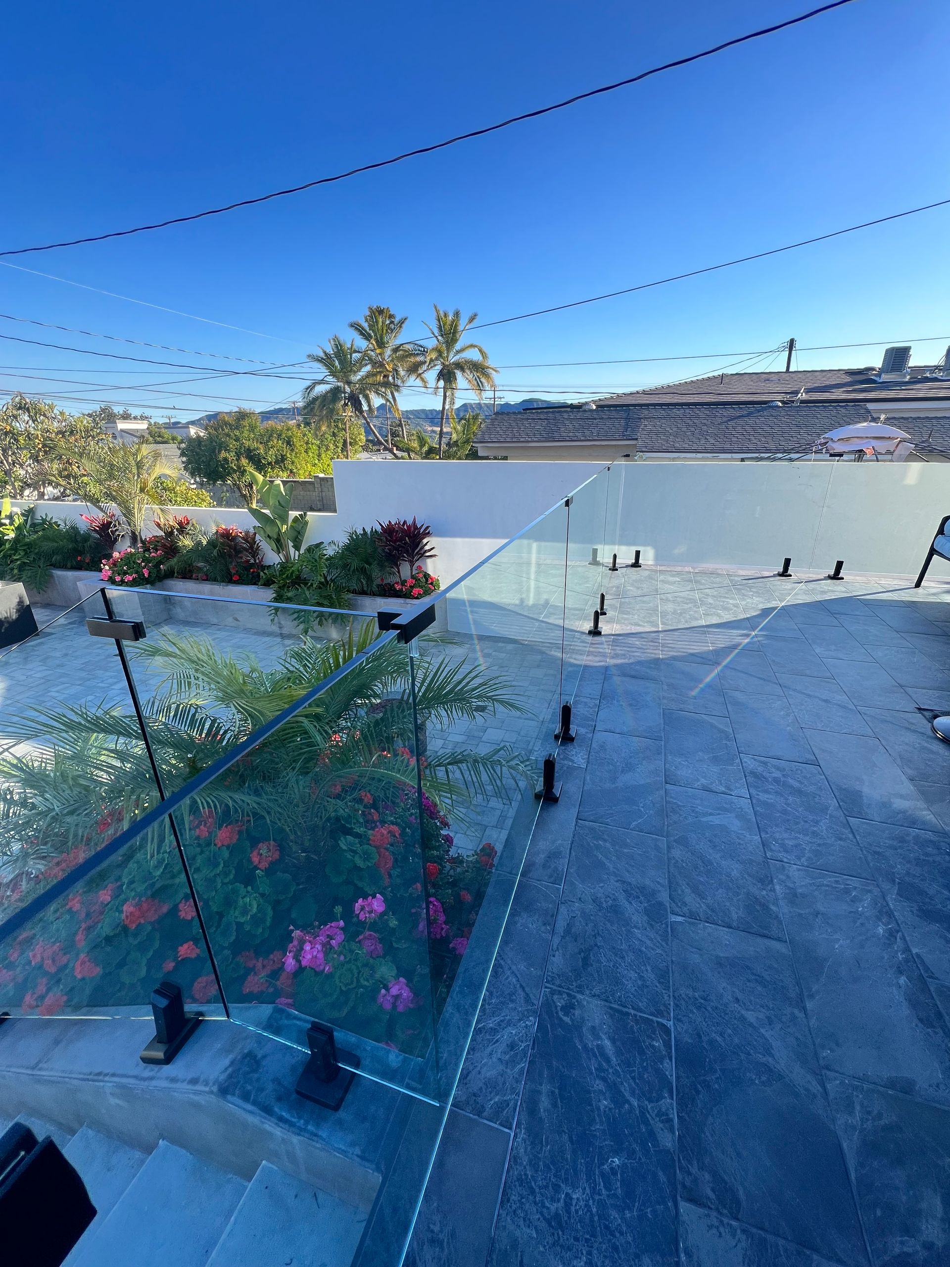 Glass railing on a stone patio overlooking a yard with flowers and palm trees, bright blue sky overhead.