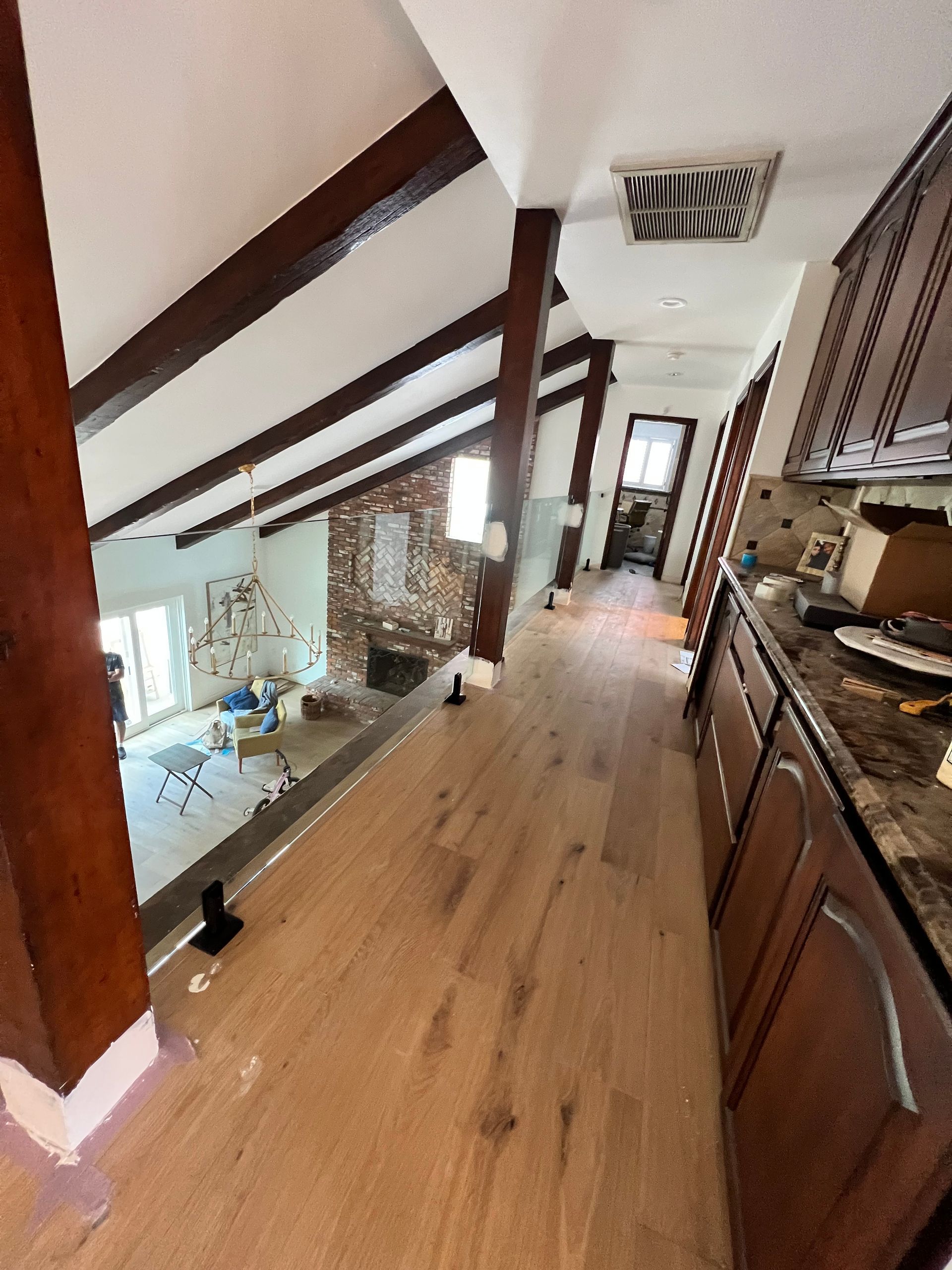 Interior with wood beams, light wood floors, kitchen cabinets, and a living area visible in the distance.