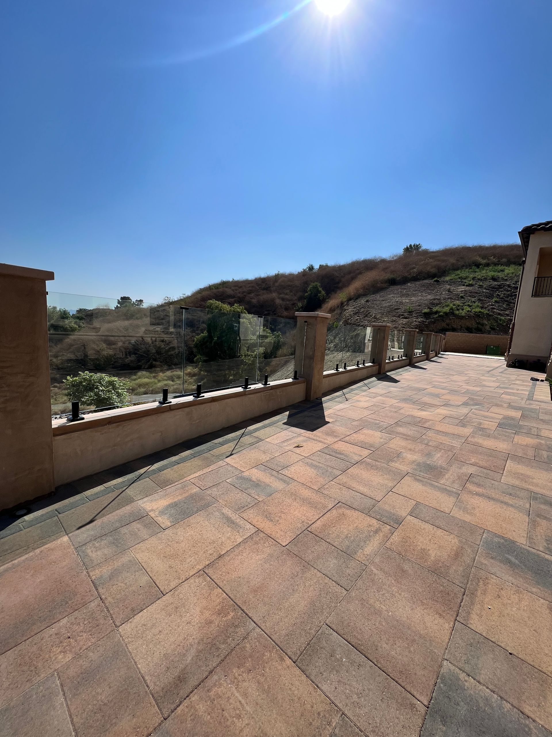 Patio with glass railing, brick pavers, and a view of dry hills under a bright blue sky.