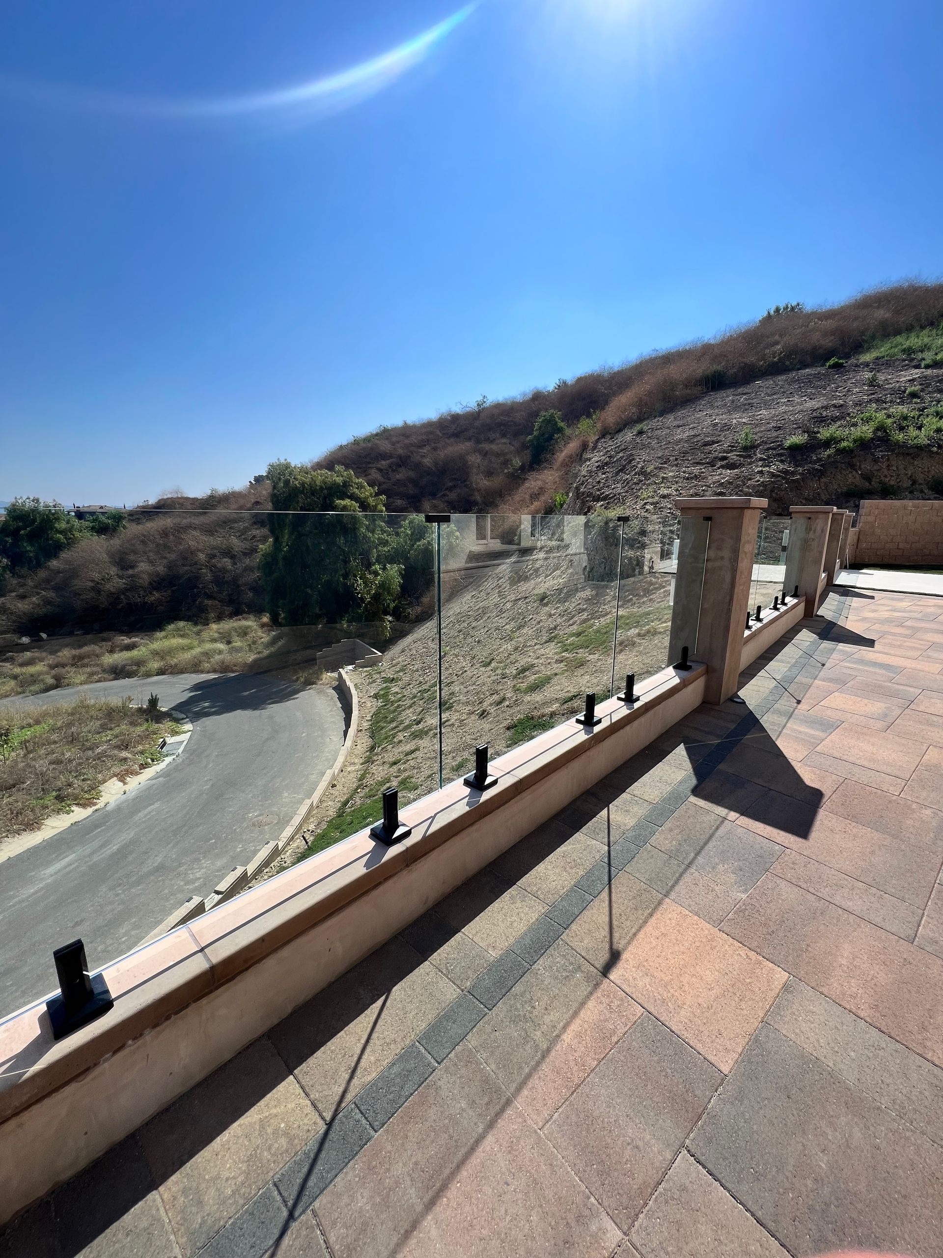 Patio overlooking a winding road and hillside with clear glass railing. Sunny day.