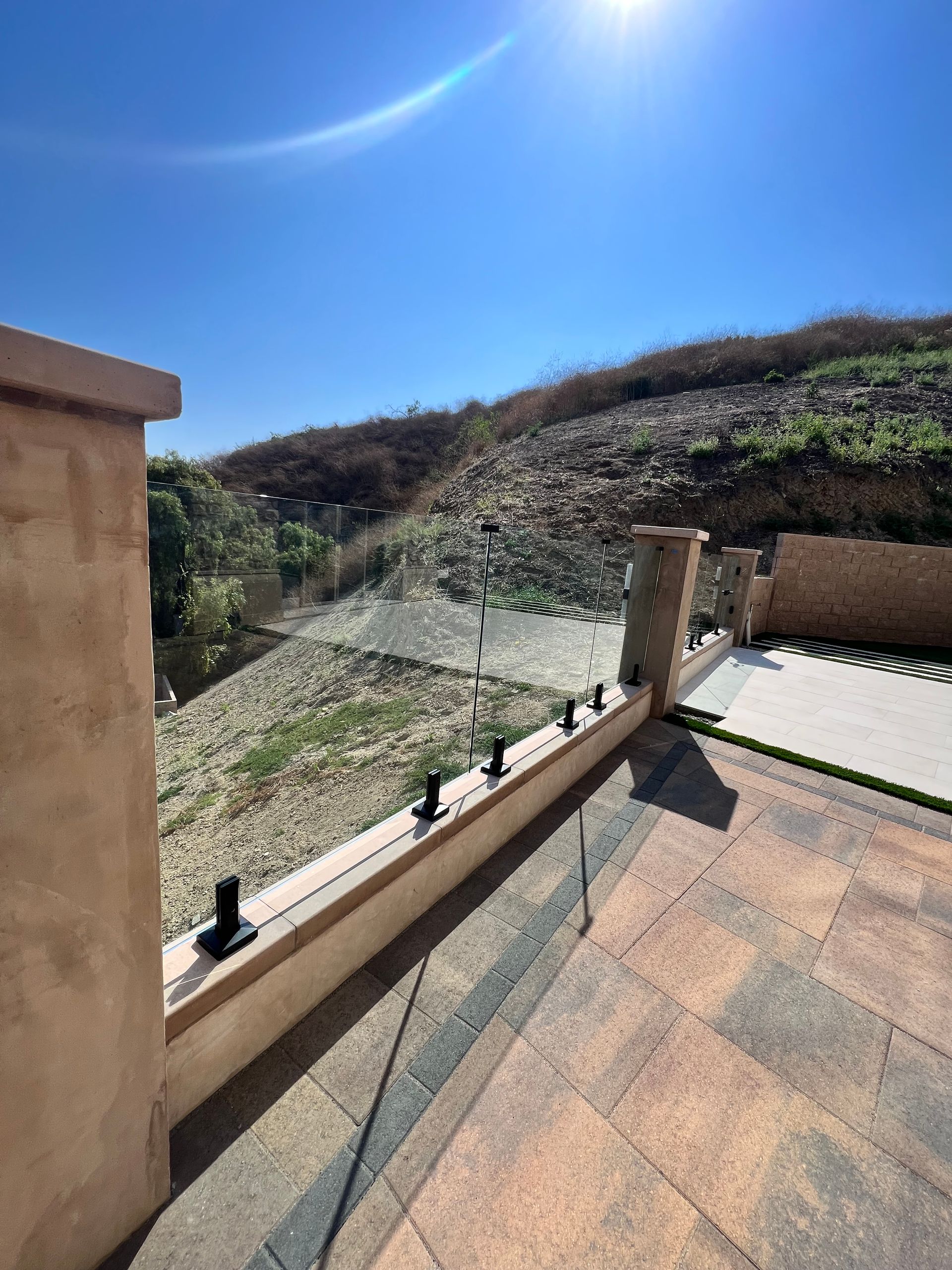 Patio with glass railing overlooking a hillside under a bright blue sky.
