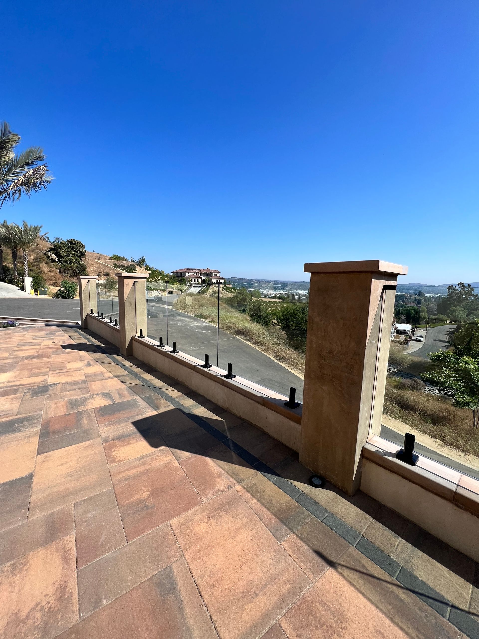 Patio with brick pavers, glass railing, and concrete posts, overlooking a sunny suburban street.